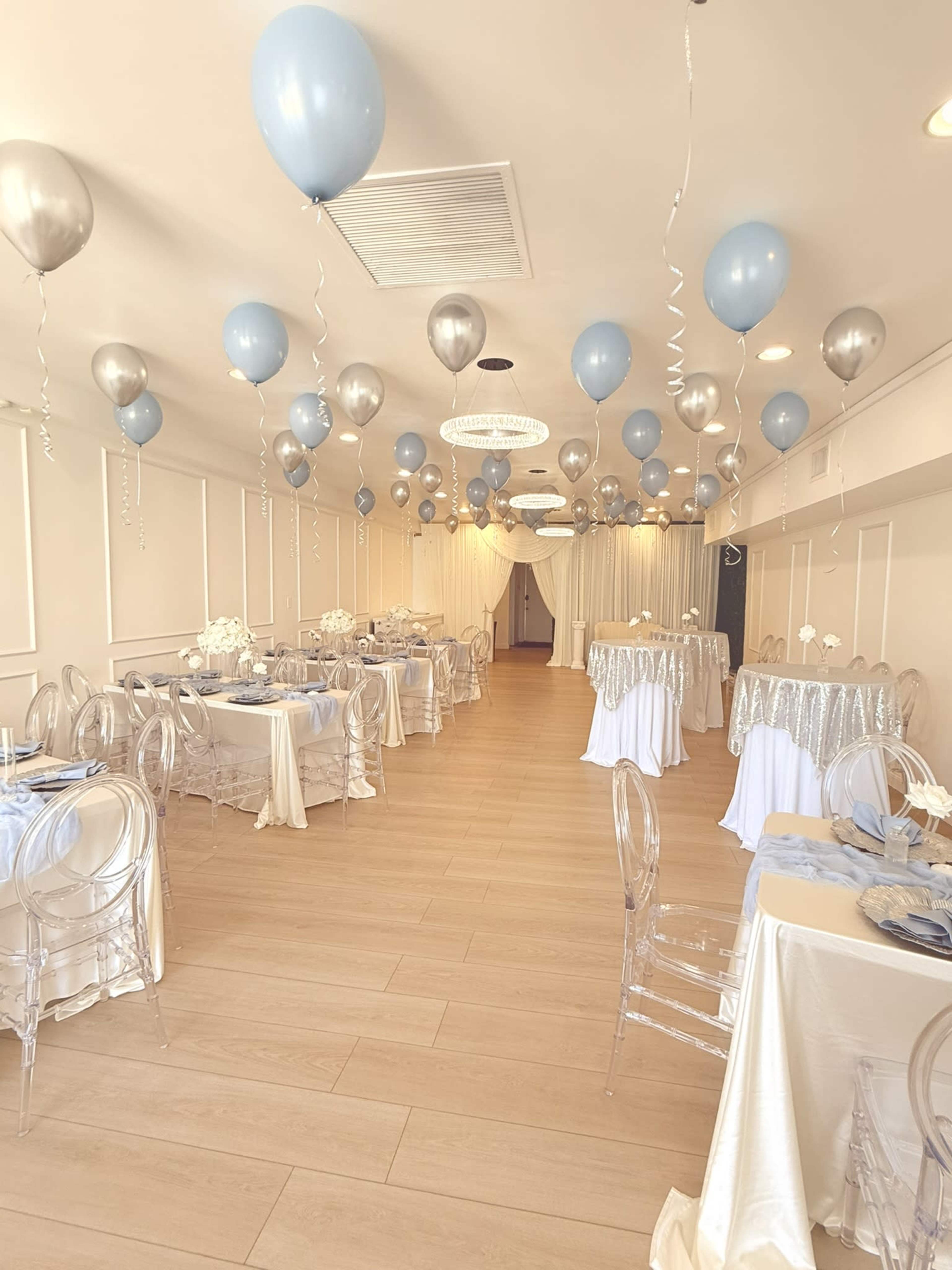 The image shows a decorated event space featuring tables draped in white and silver tablecloths, clear chairs, and blue and silver balloons suspended from the ceiling.