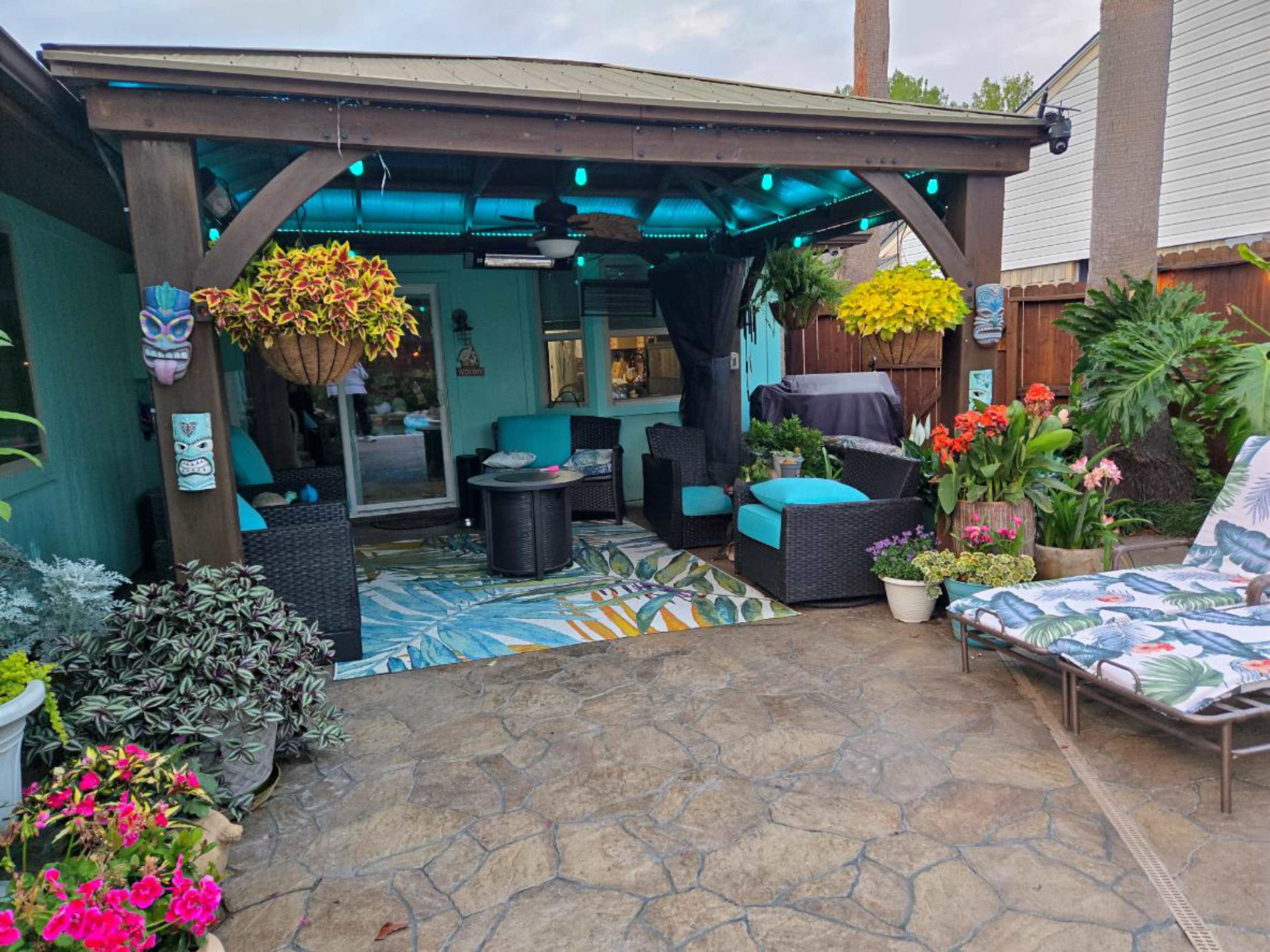 The image shows a patio area with a covered seating arrangement, colorful plants, and decorative lights.
