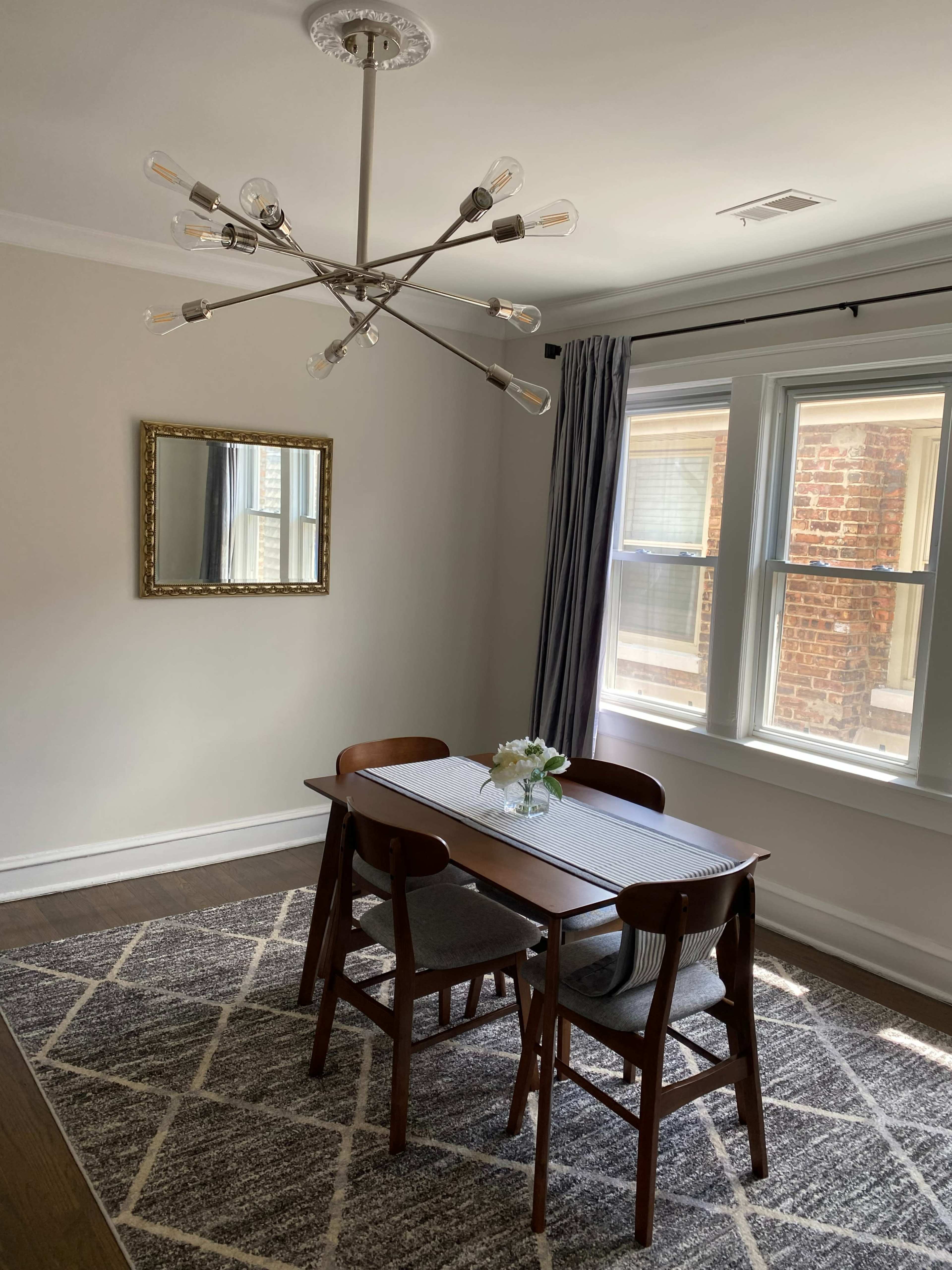 A dining table with four chairs is set in a light-filled room featuring a mirror and a modern chandelier.