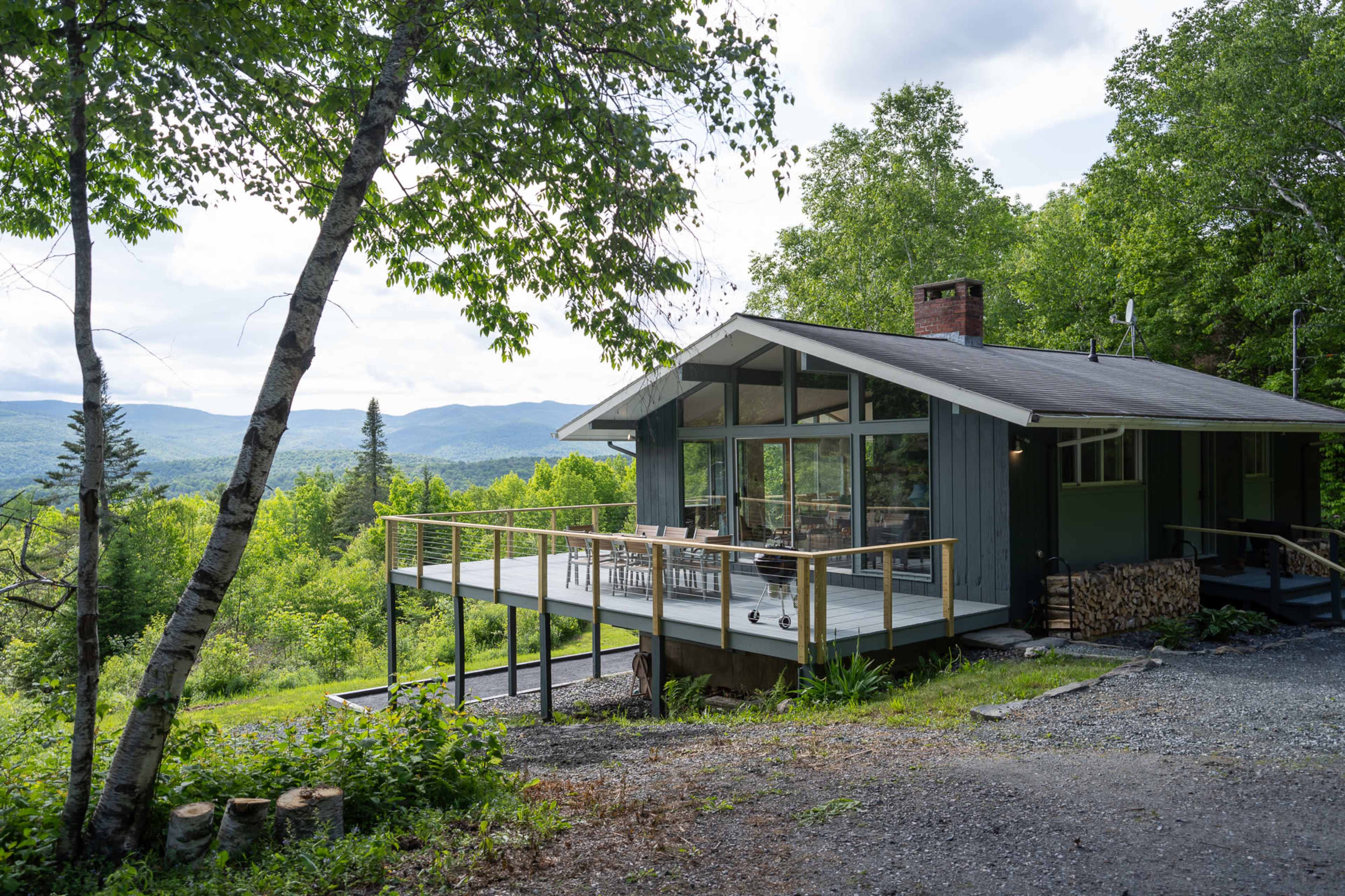 A modern cabin with a large deck overlooks a lush green landscape and distant mountains.