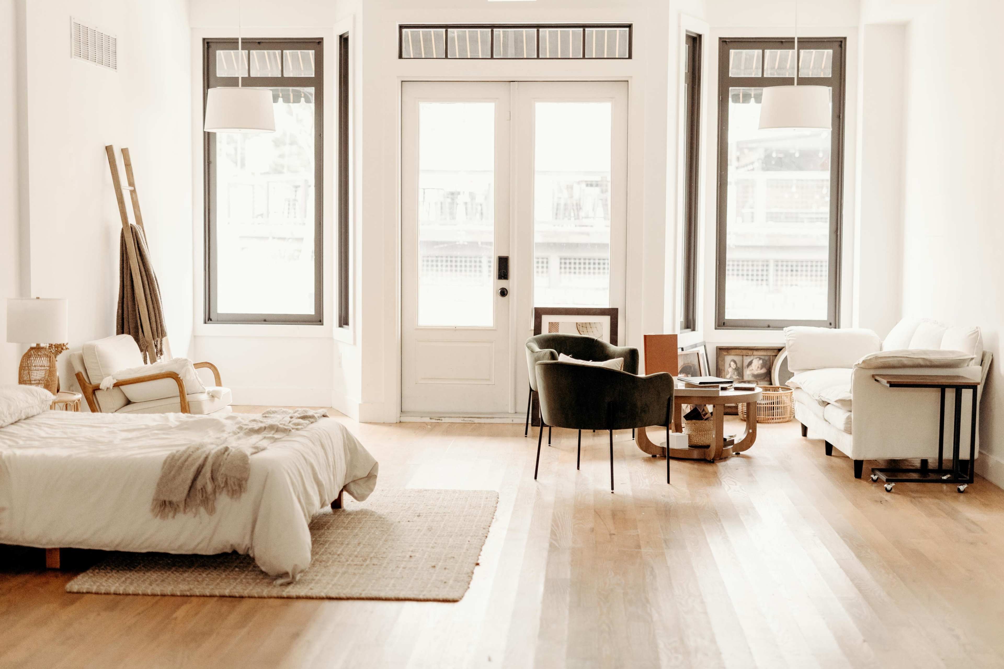 The image shows a bright, minimalist living space featuring a bed, two chairs, a small coffee table, and large windows with natural light.