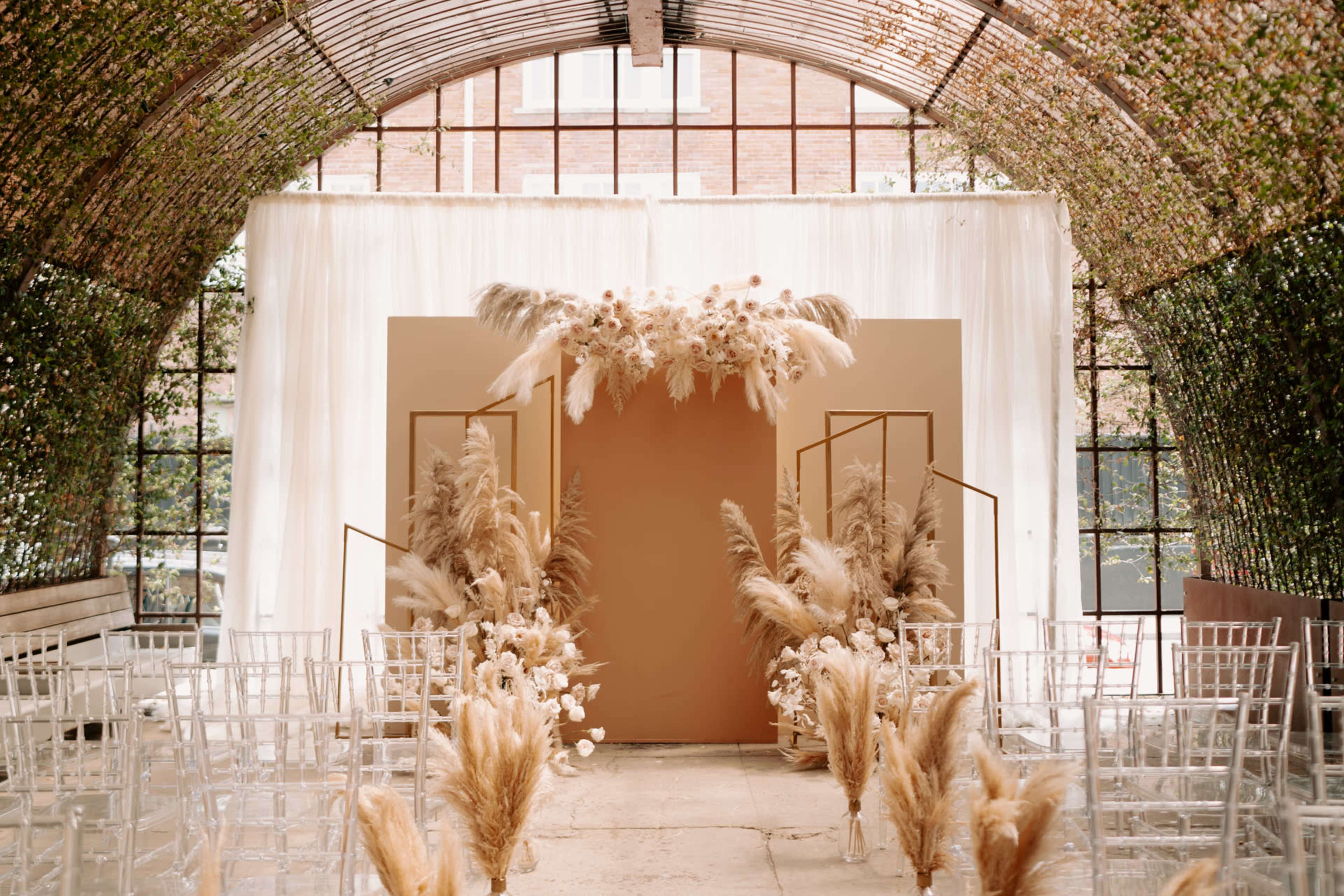 A wedding ceremony setup features an arch adorned with dried flowers and grasses, flanked by transparent chairs and a backdrop of greenery.