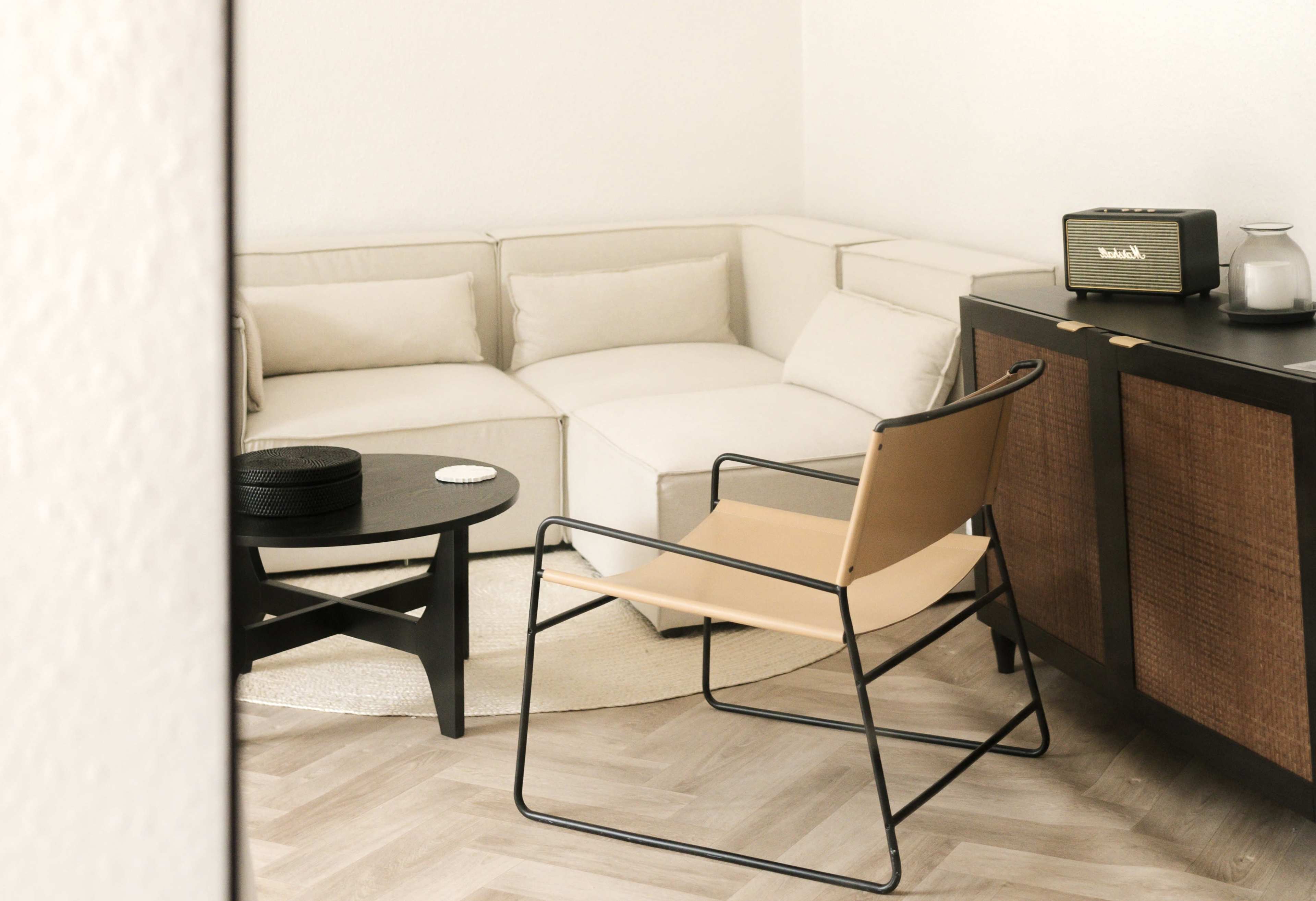A modern living room features a sectional sofa, a round black table, a light-colored chair, and a vintage radio set against a textured cabinet.