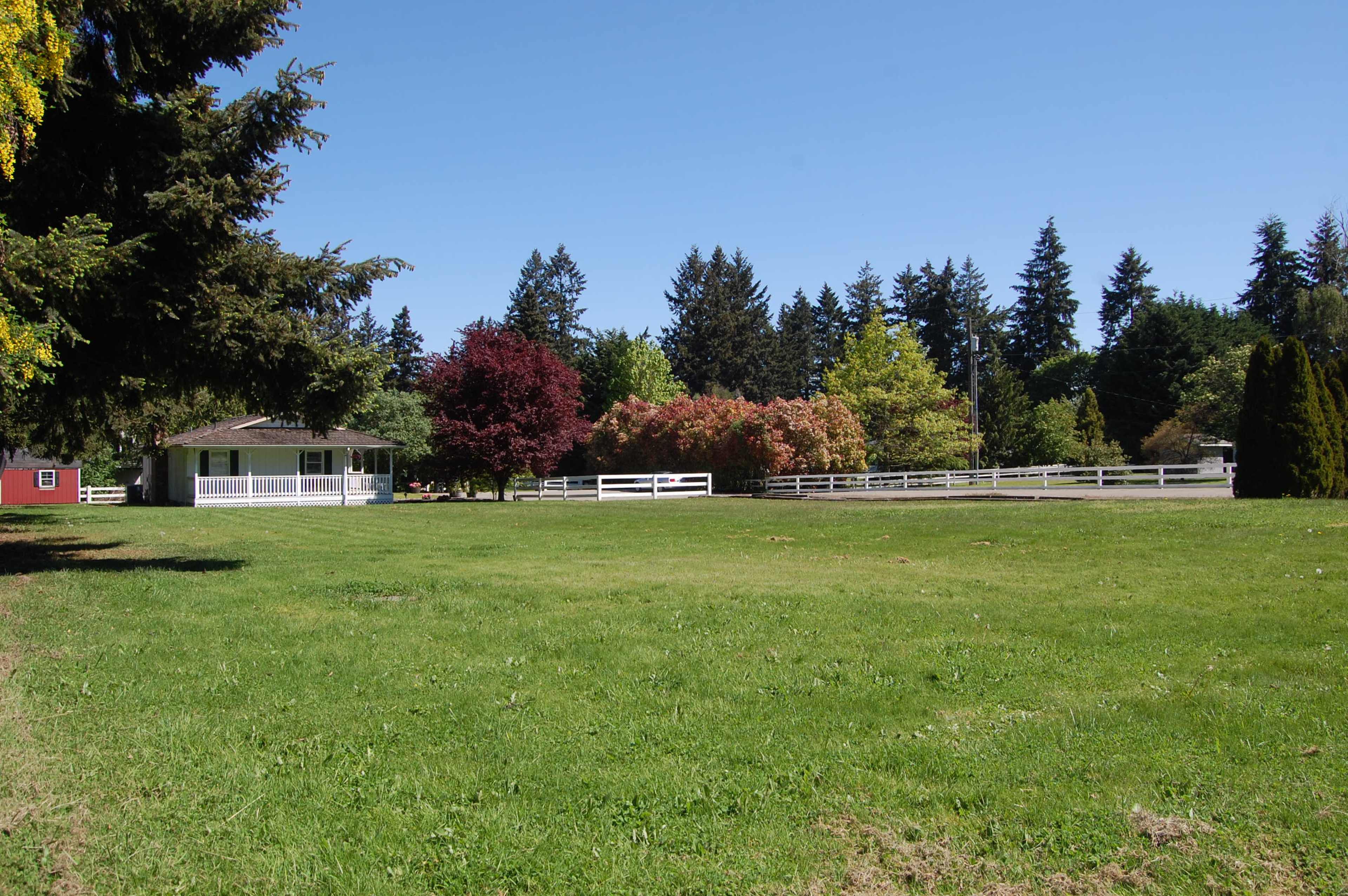 The image shows a spacious green field with a farmhouse and several trees in a rural setting.