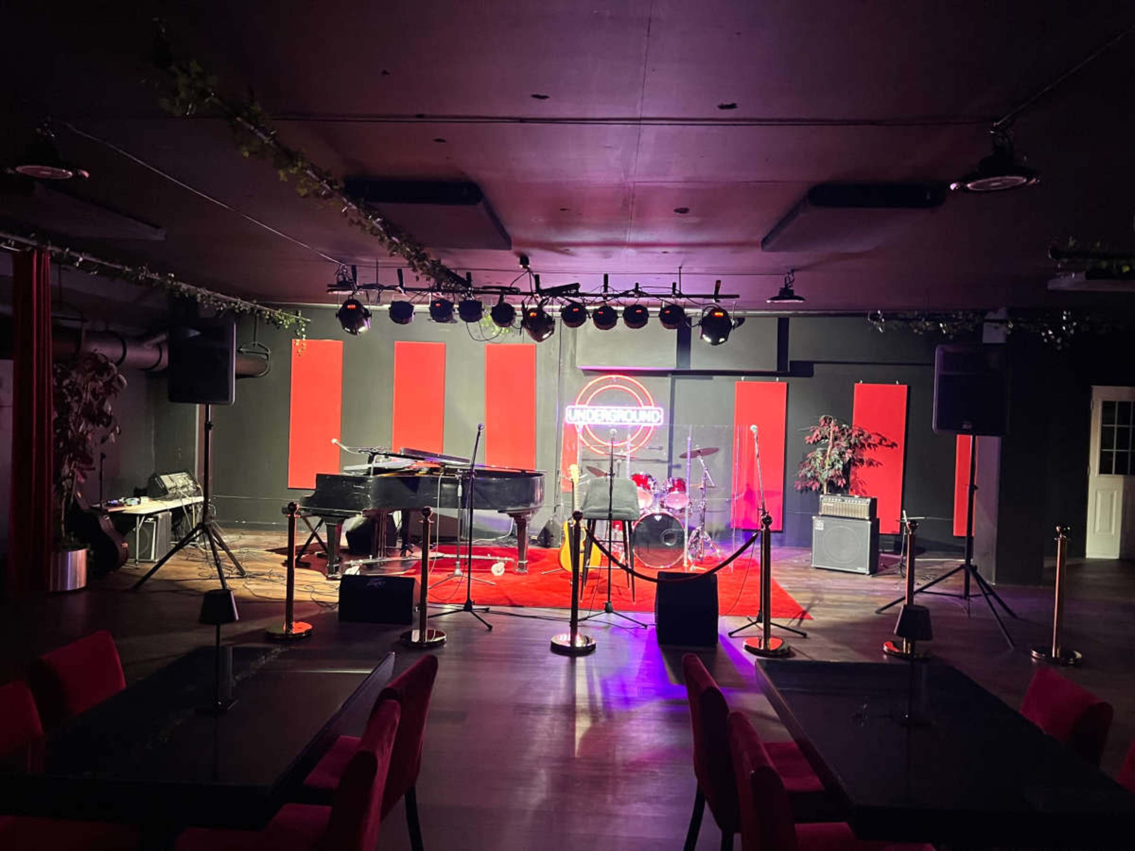 The image shows a small stage setup in a dimly lit venue, featuring a piano, microphone, and drum set, surrounded by red accent walls and tables arranged in front.