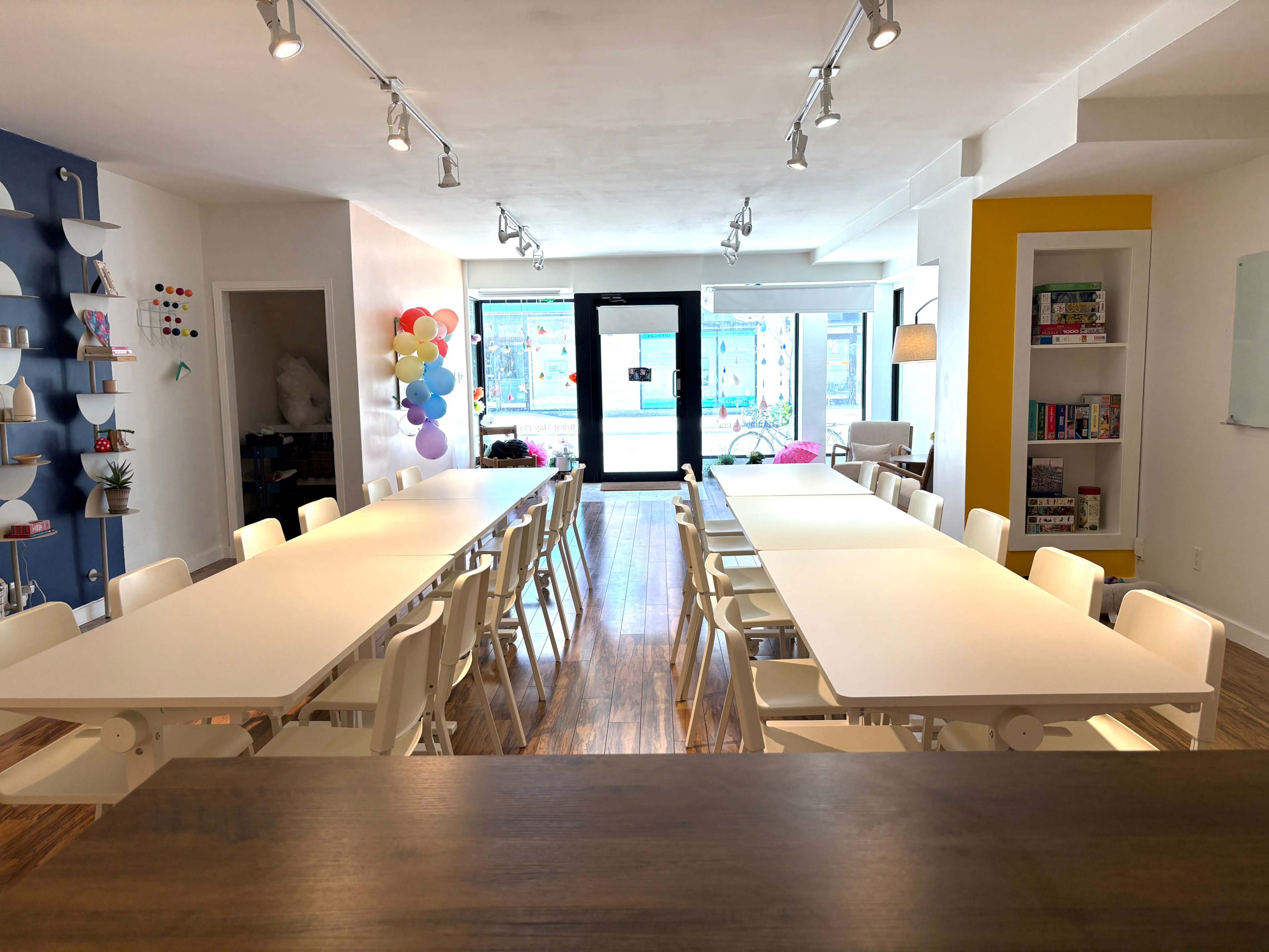 A spacious room features several long tables arranged in a central layout, surrounded by white chairs, with large windows allowing natural light and colorful decor on the walls.