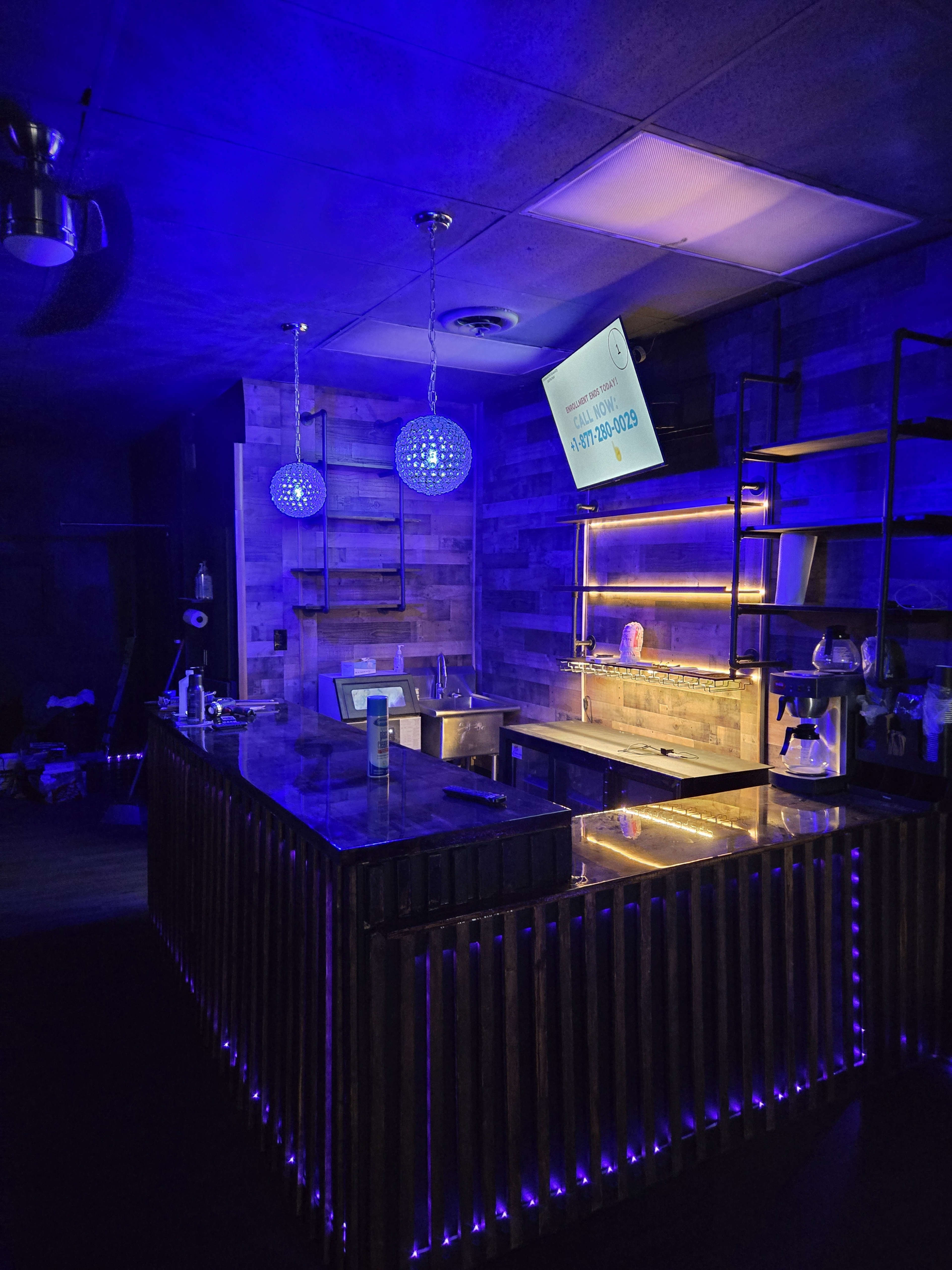 The image shows a modern bar area with a wooden counter, illuminated by blue and yellow lights, and decorative glass fixtures hanging above.