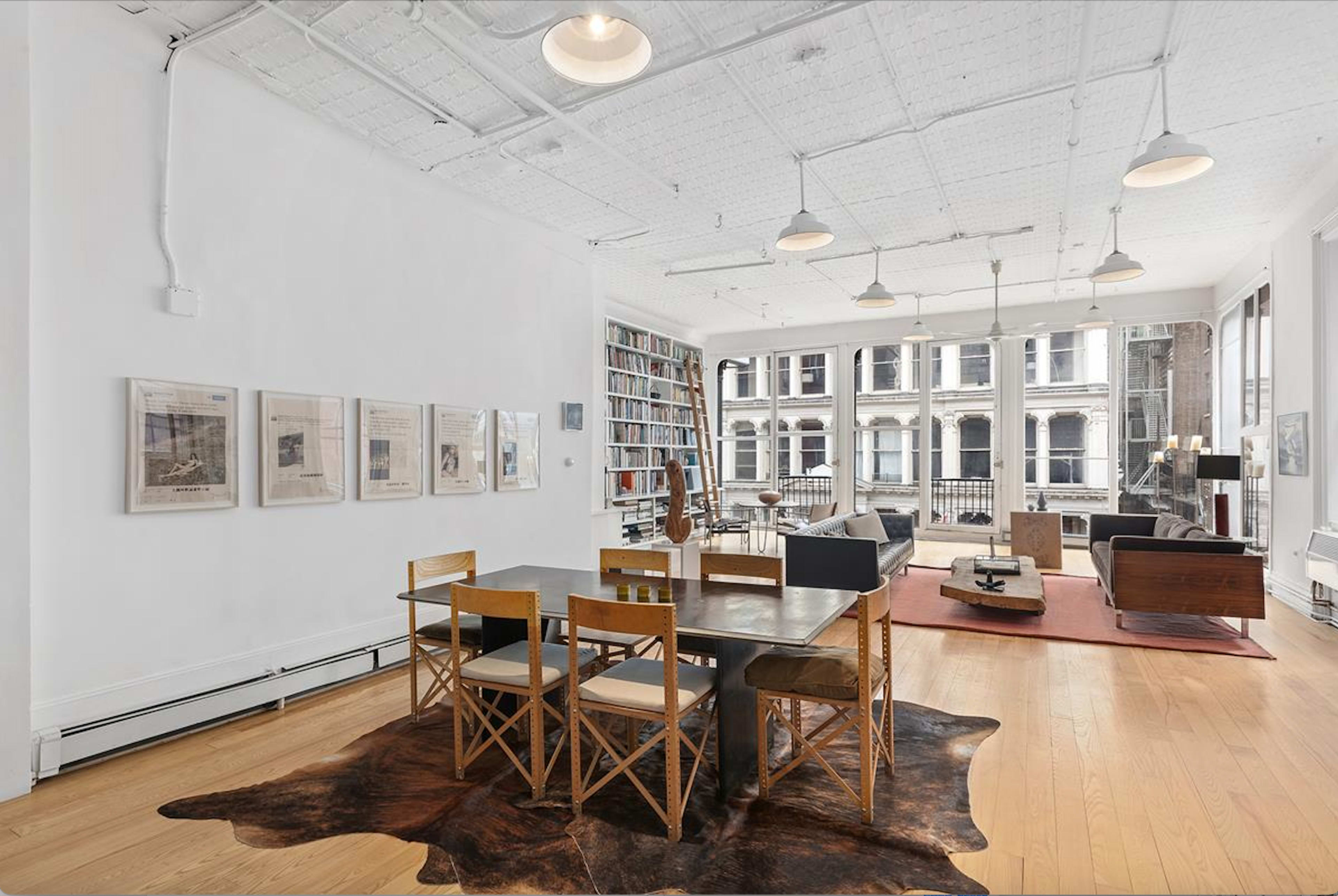 A modern, open living space with a dining area featuring a black table and wooden chairs, large windows overlooking a cityscape, and a collection of bookshelves against the wall.