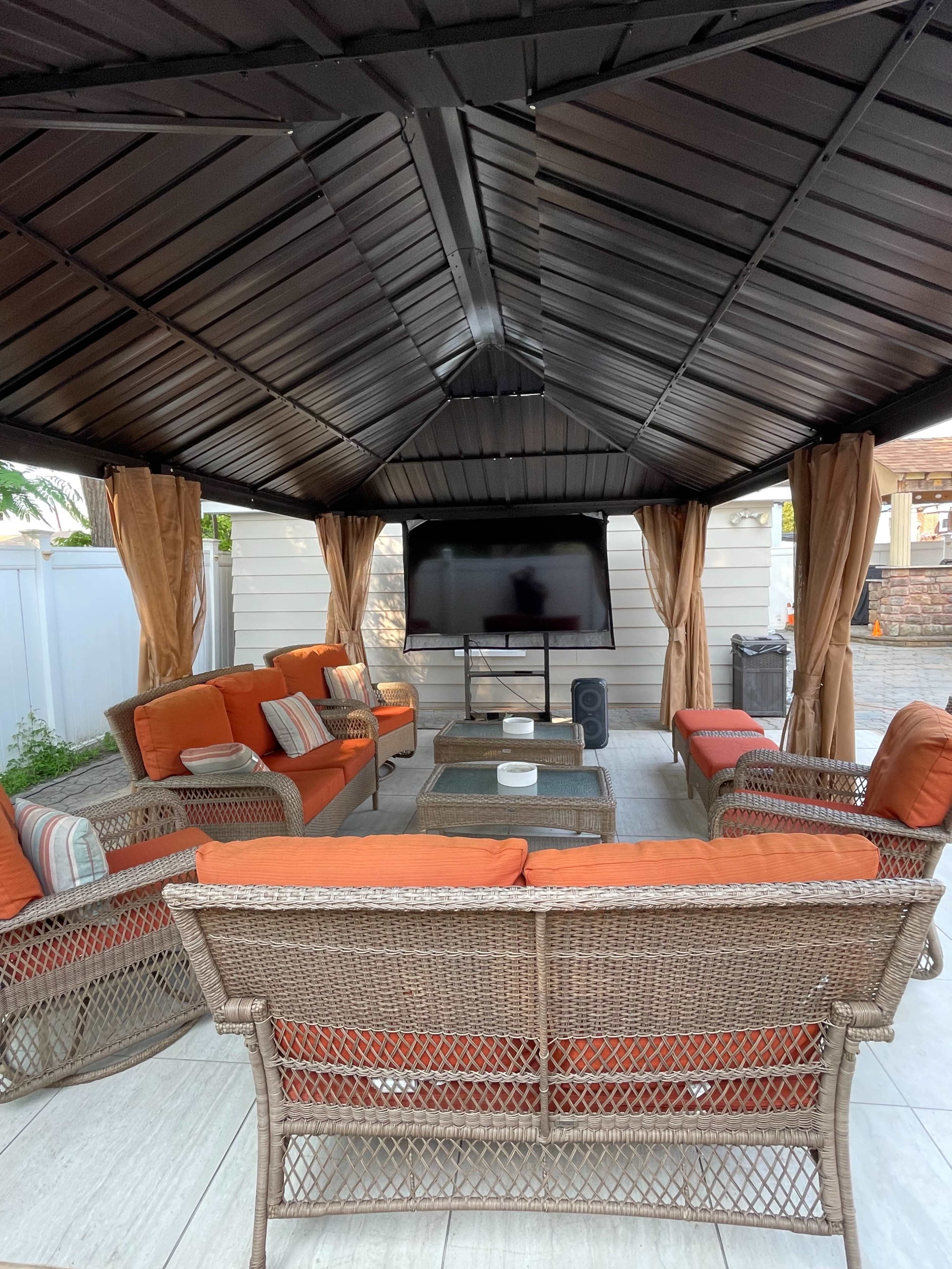 A covered outdoor seating area features orange cushions on wicker furniture, with a television mounted on the wall and curtains along the sides.