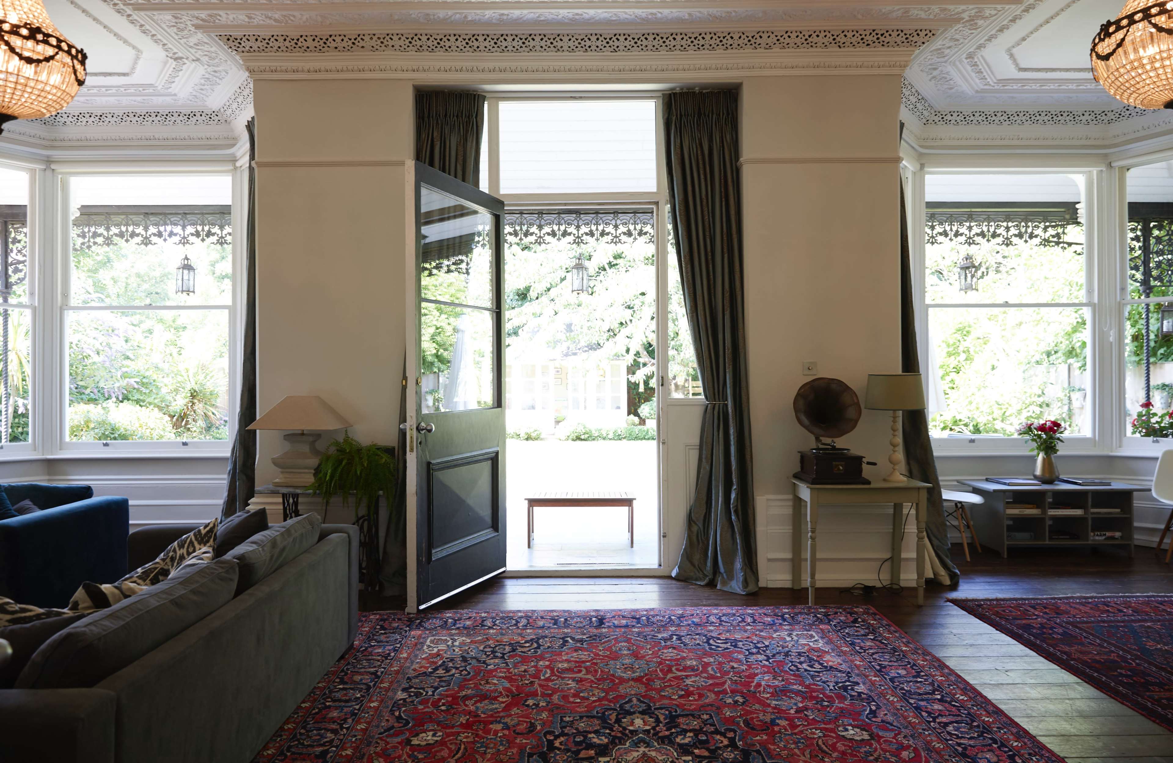 A spacious living room features a large open door leading to a garden, with a plush sofa, a vintage record player, and decorative curtains near the windows.