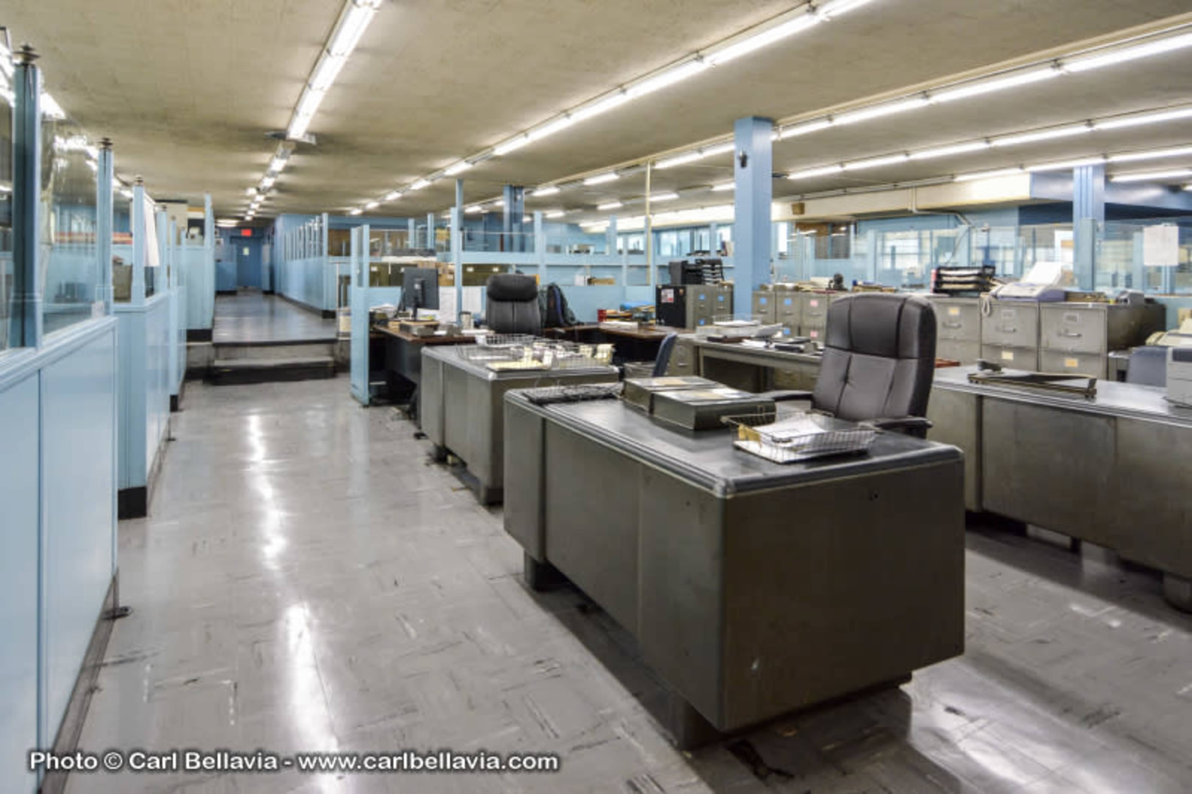 A spacious, vacant office area with metal desks and filing cabinets lined along the sides, illuminated by fluorescent lights overhead.