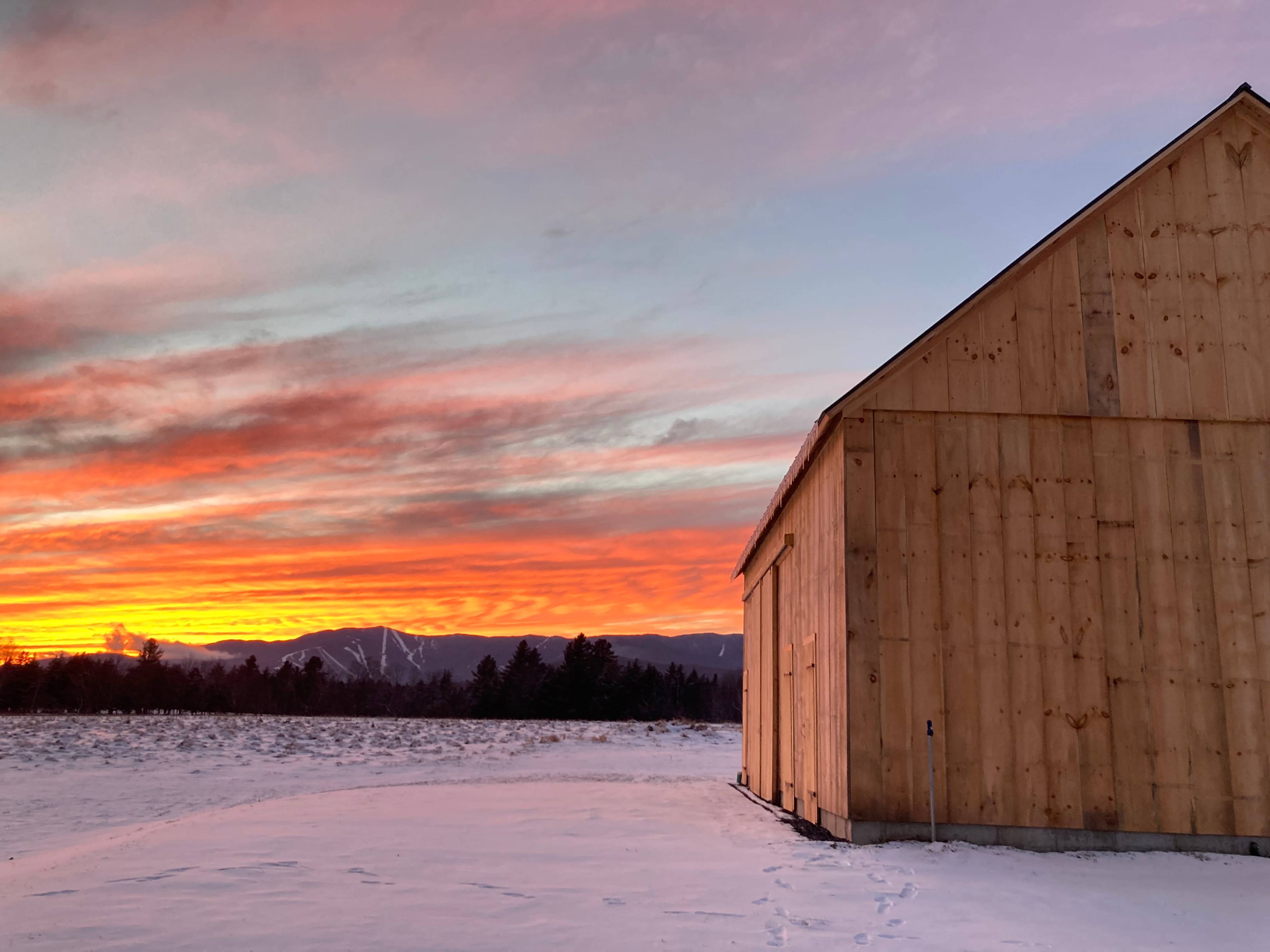 A wooden barn stands against a vibrant sunset with streaks of orange and pink in the sky, framed by a snowy field and distant mountains.