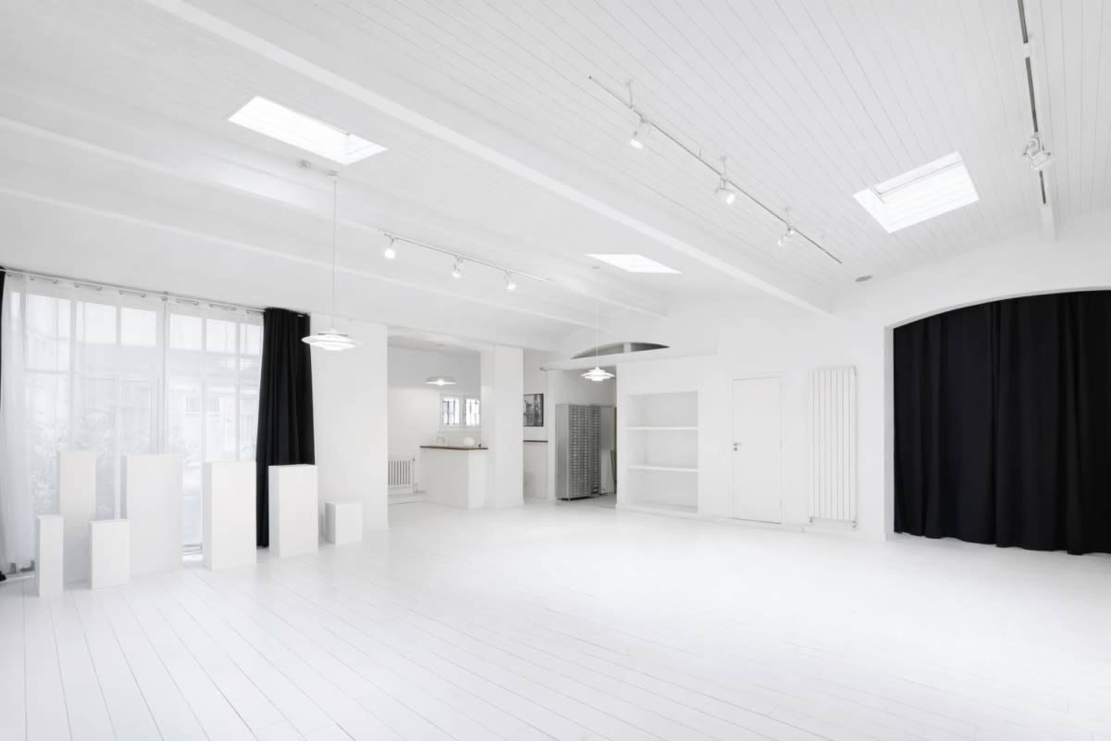 The image shows a spacious white room with high ceilings, wooden floors, and minimal furnishings, featuring large windows and two black curtains.