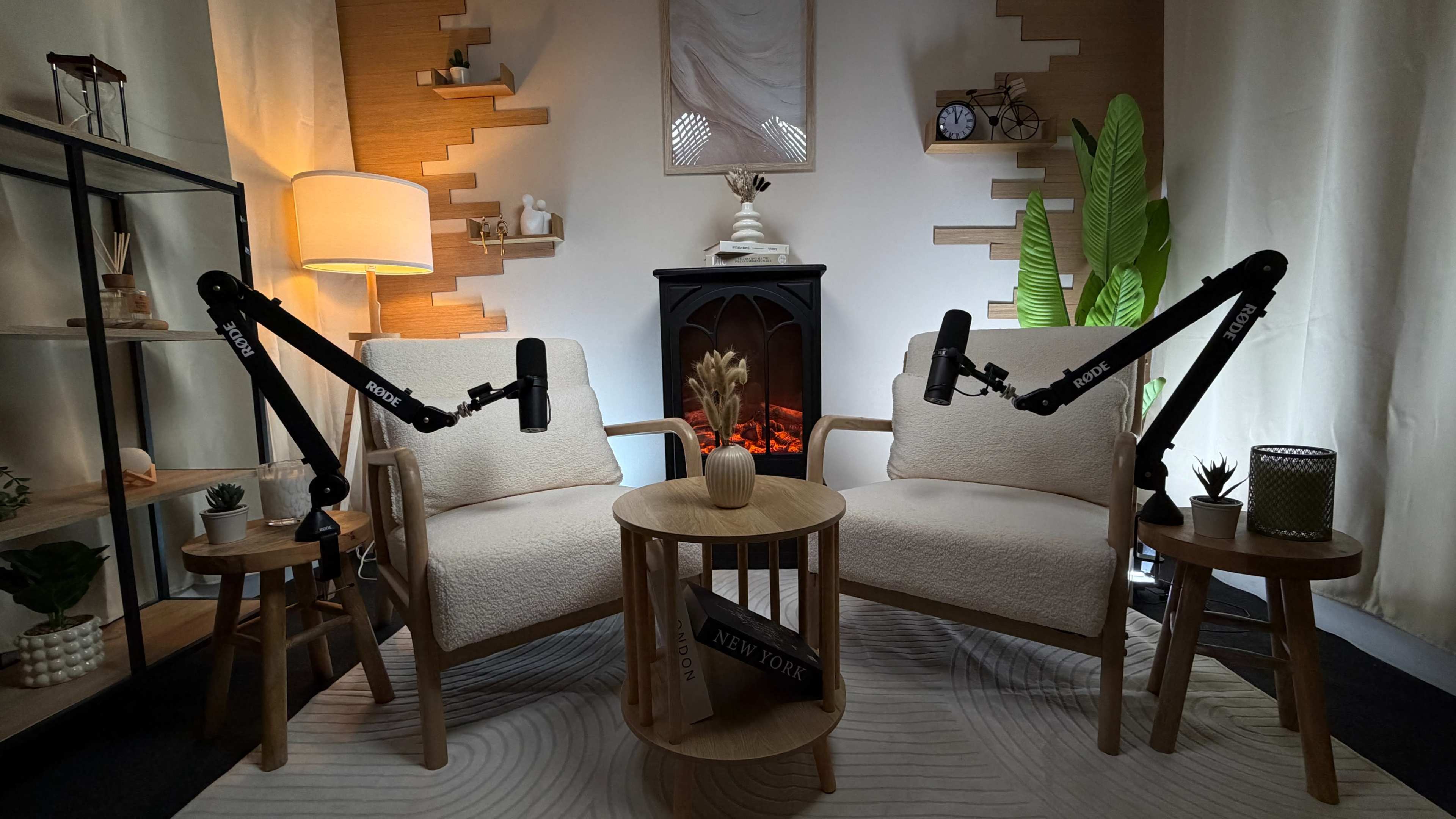 The image shows a cozy indoor setup featuring two chairs with microphones mounted on stands, a small wooden table, and decorative plants against a warm background.