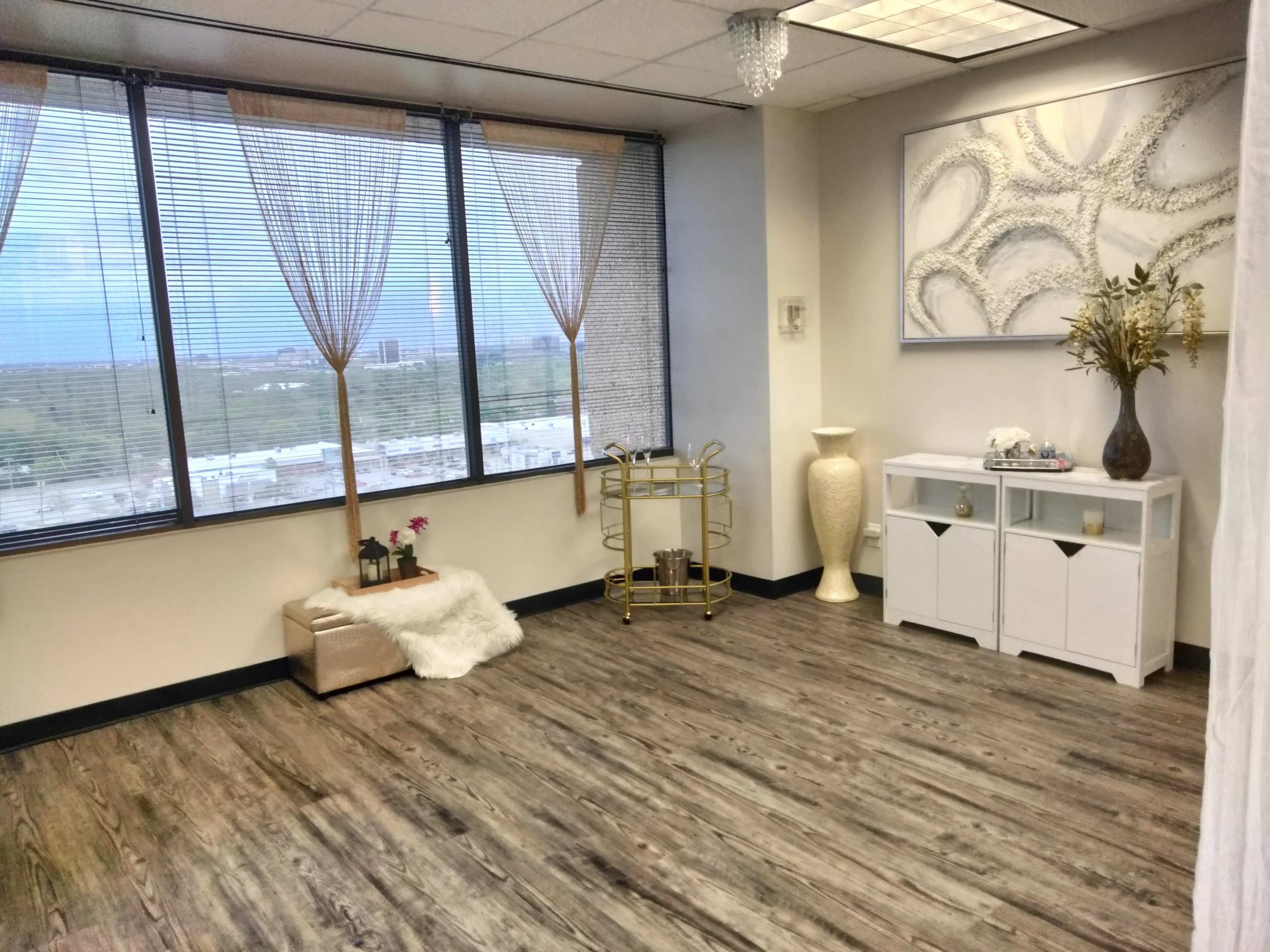 A spacious room with large windows, wooden flooring, and minimalistic furniture, including a gold cart and a white cabinet.