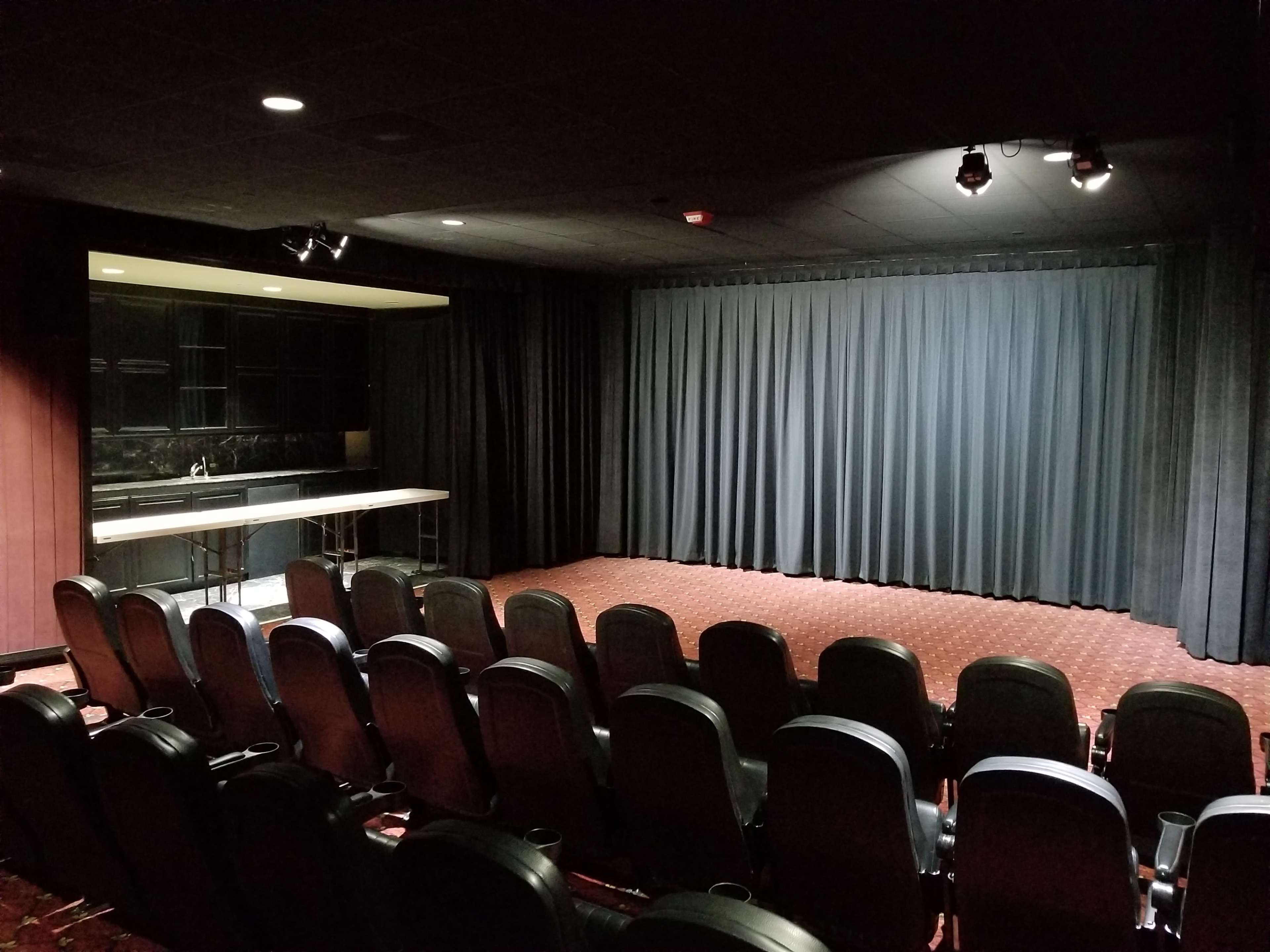 The image shows a small theater with rows of black chairs facing a stage covered by a curtain, and a counter area on the left.