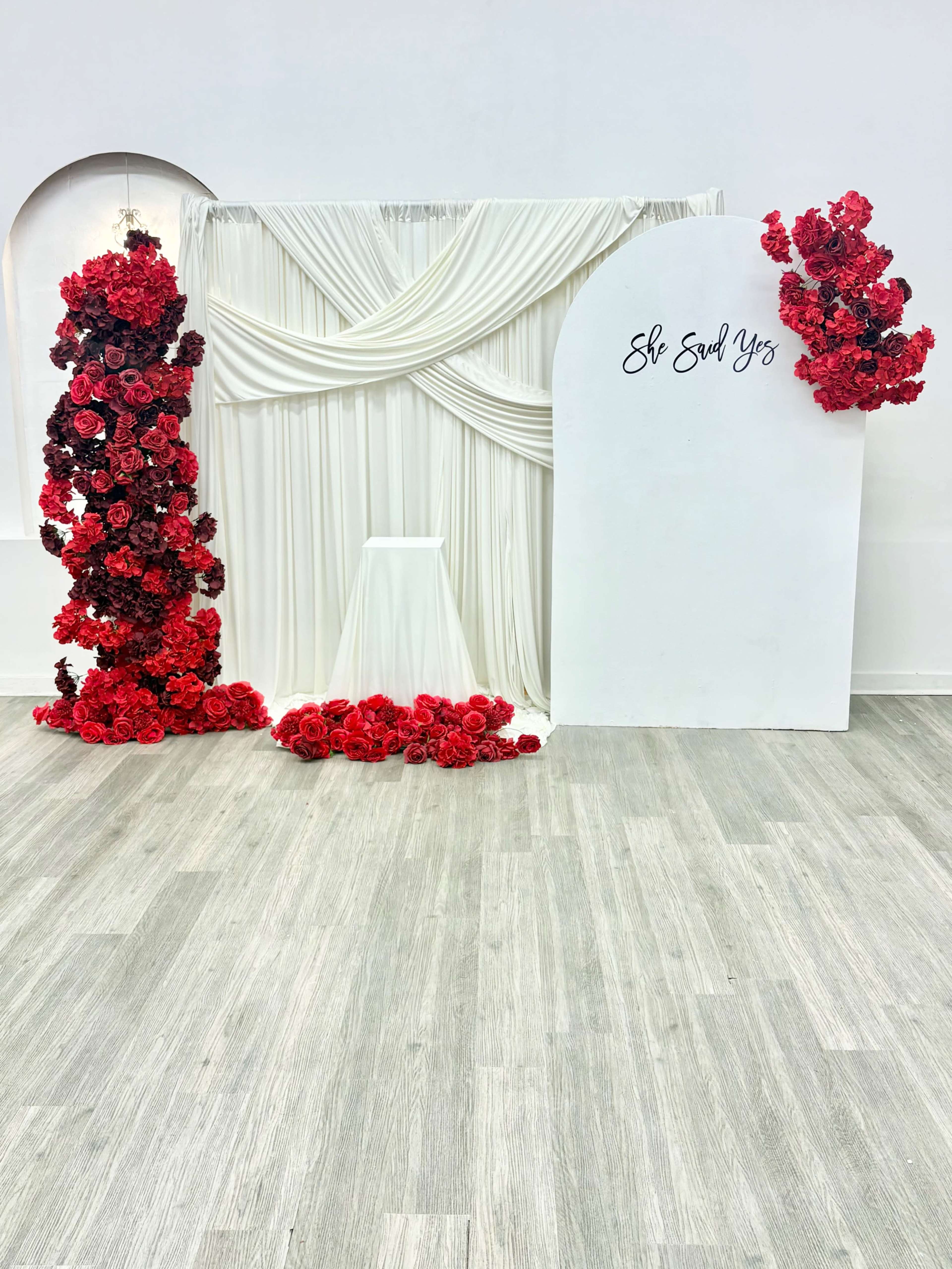 The image features a decorative backdrop with cascading white drapes and red floral arrangements, along with a white sign that reads "She Said Yes."