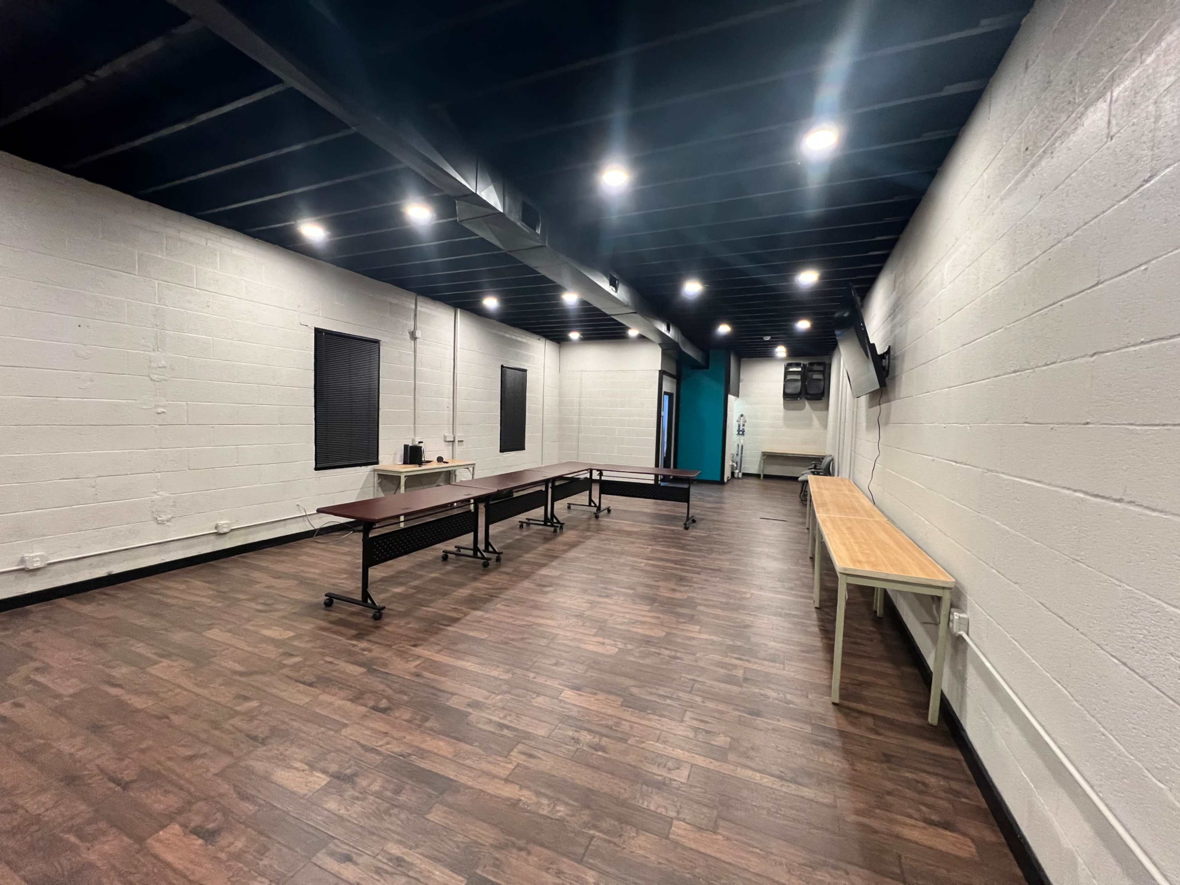 A spacious room with wooden floors, several tables, and a few windows, illuminated by overhead lights.
