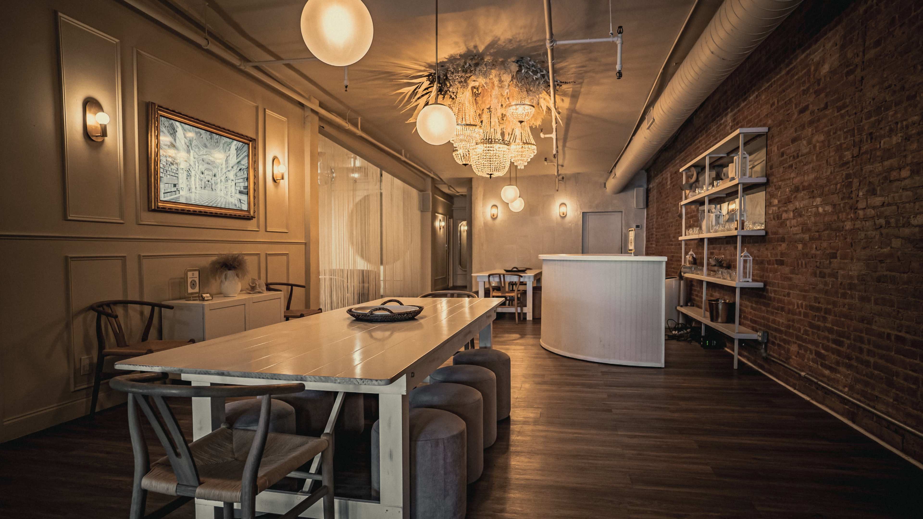 The image showcases a modern interior space featuring a long dining table, various seating options, a bar area, and decorative lighting against exposed brick walls.