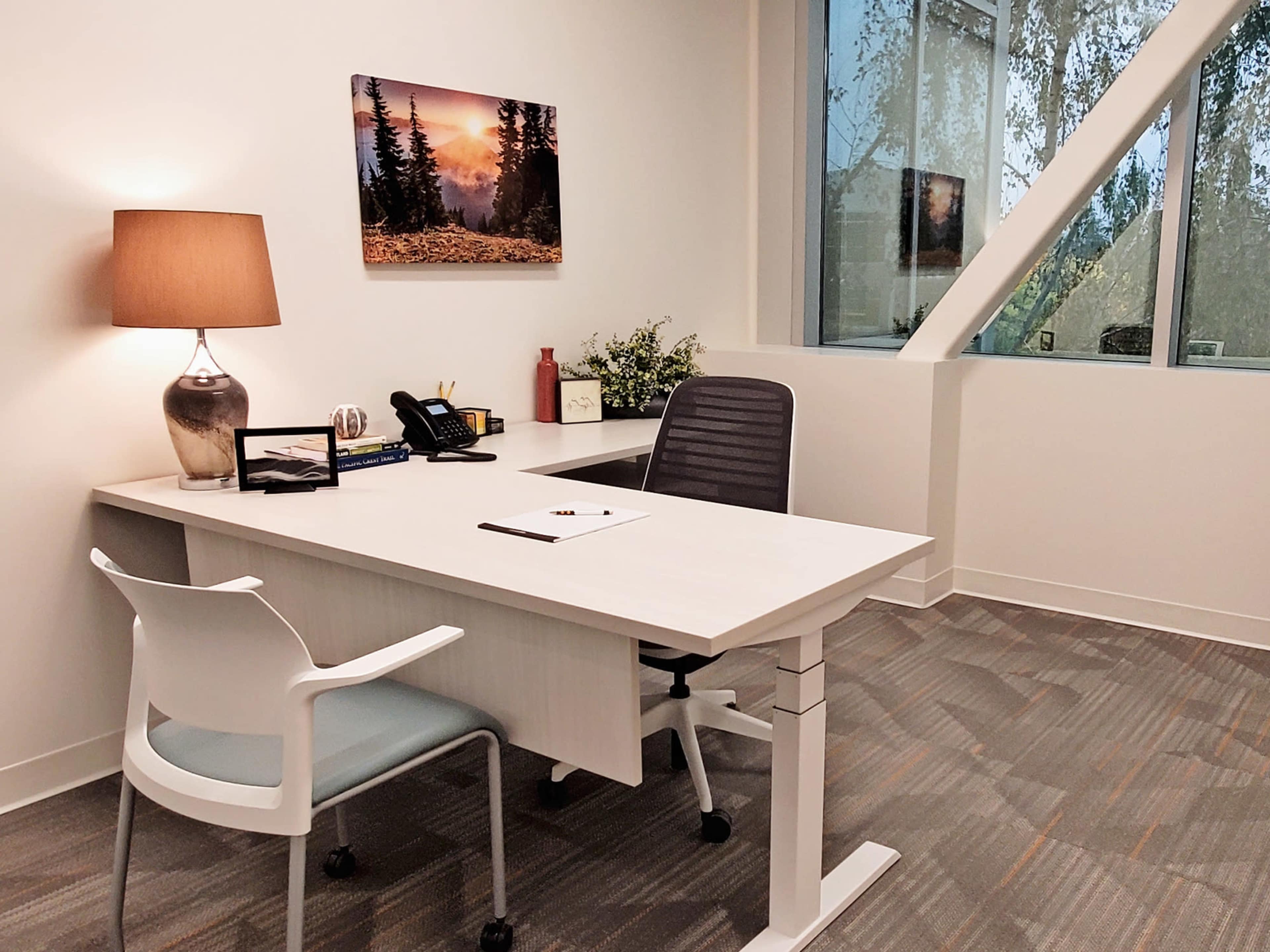 A modern office space featuring a desk, a chair, a lamp, and large windows with natural light.
