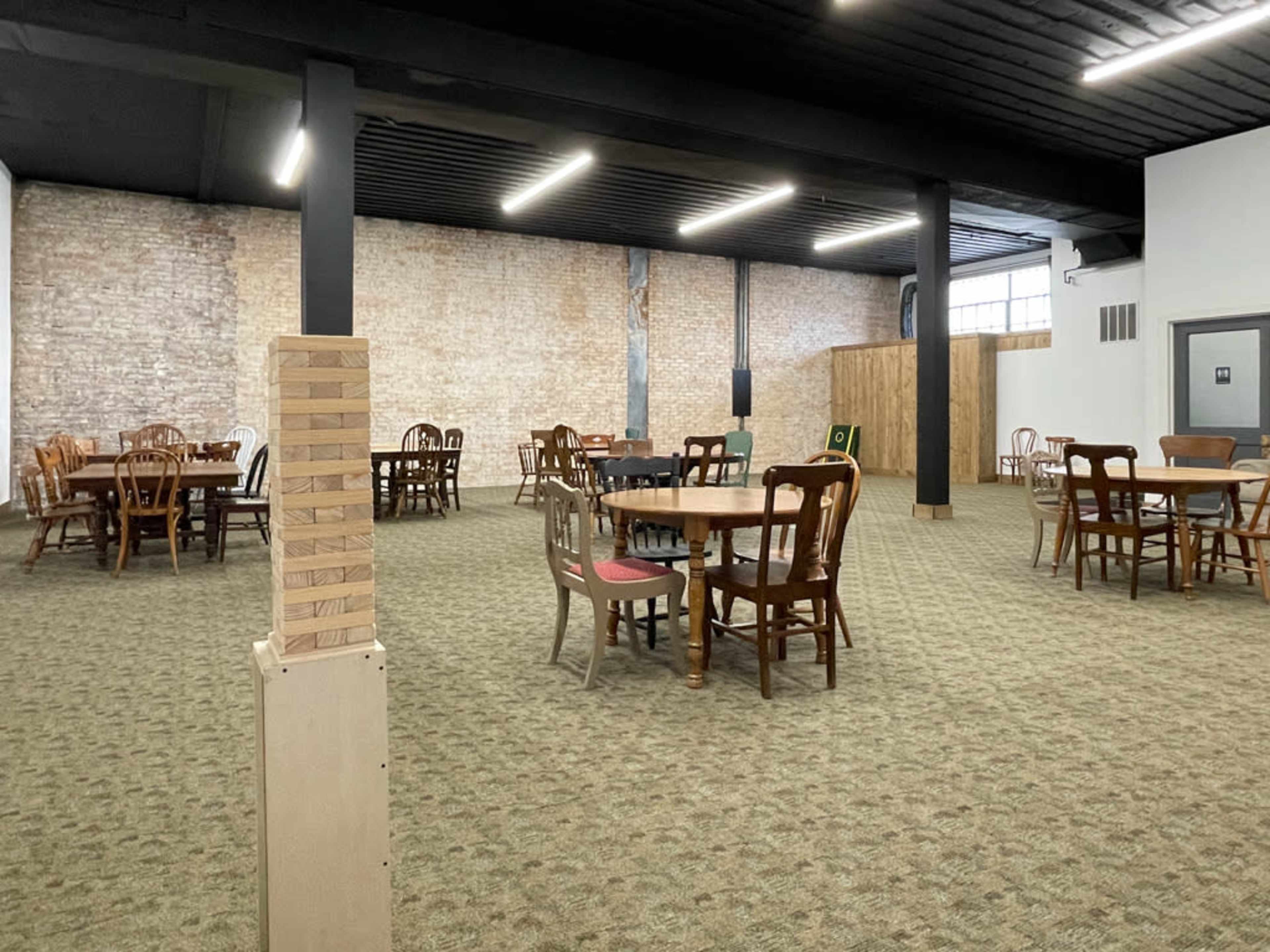 The image shows an empty room with several wooden tables and chairs arranged on a carpeted floor, with exposed brick walls and overhead lighting.
