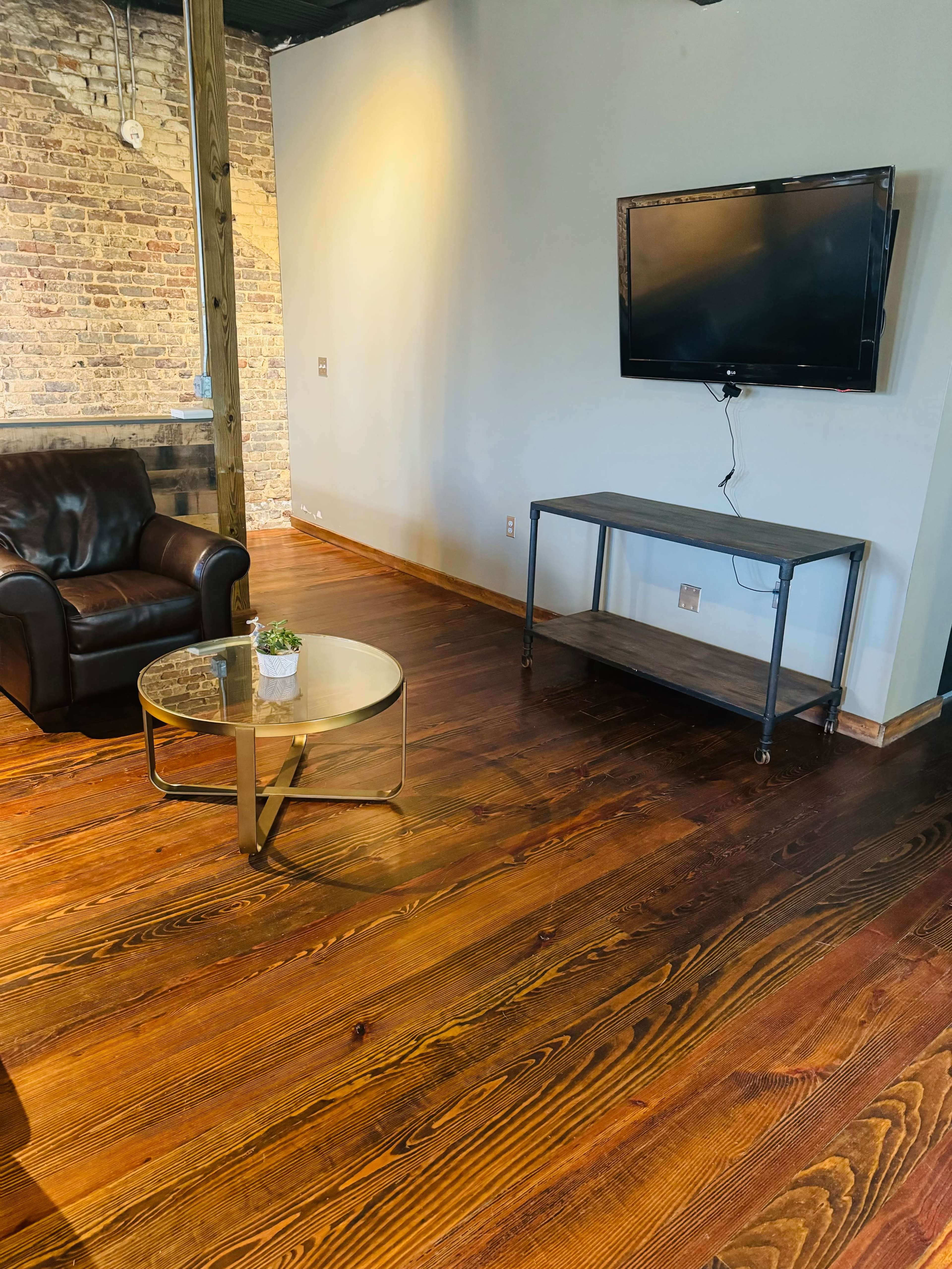 A spacious room with a leather sofa, a glass coffee table, and a television mounted on a wall above a metal media stand, featuring wooden floors and exposed brick walls.