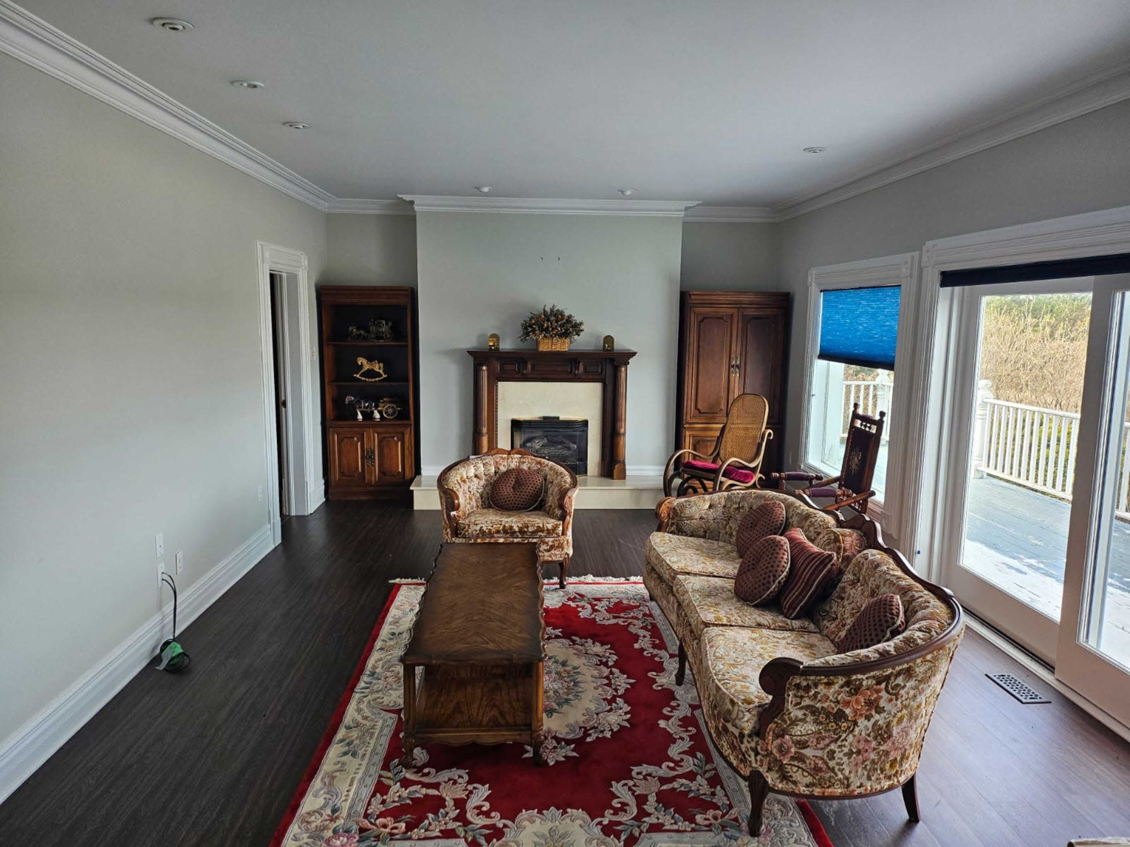 A living room with floral-patterned couches, a wooden coffee table, a fireplace flanked by wooden cabinets, and large windows with blue blinds.