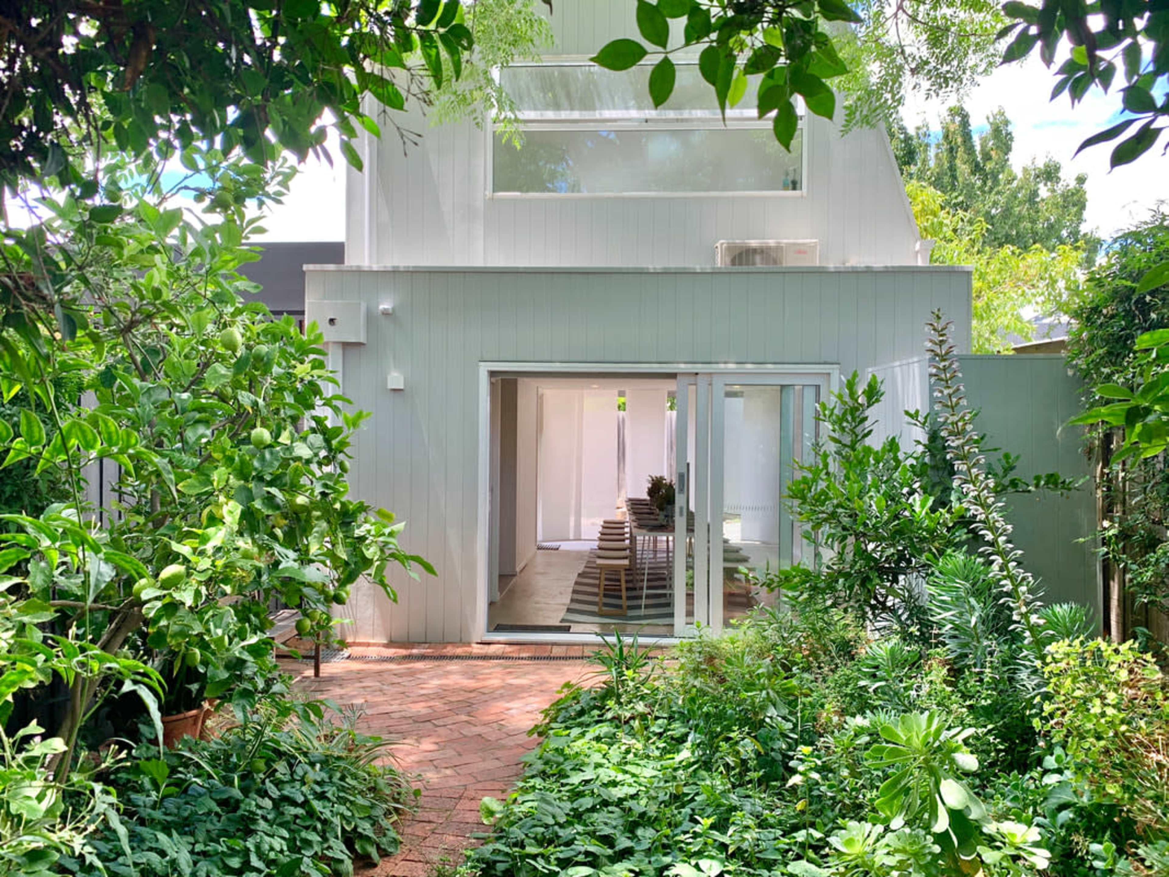 A modern, two-story house with large glass doors is surrounded by greenery and a brick pathway.