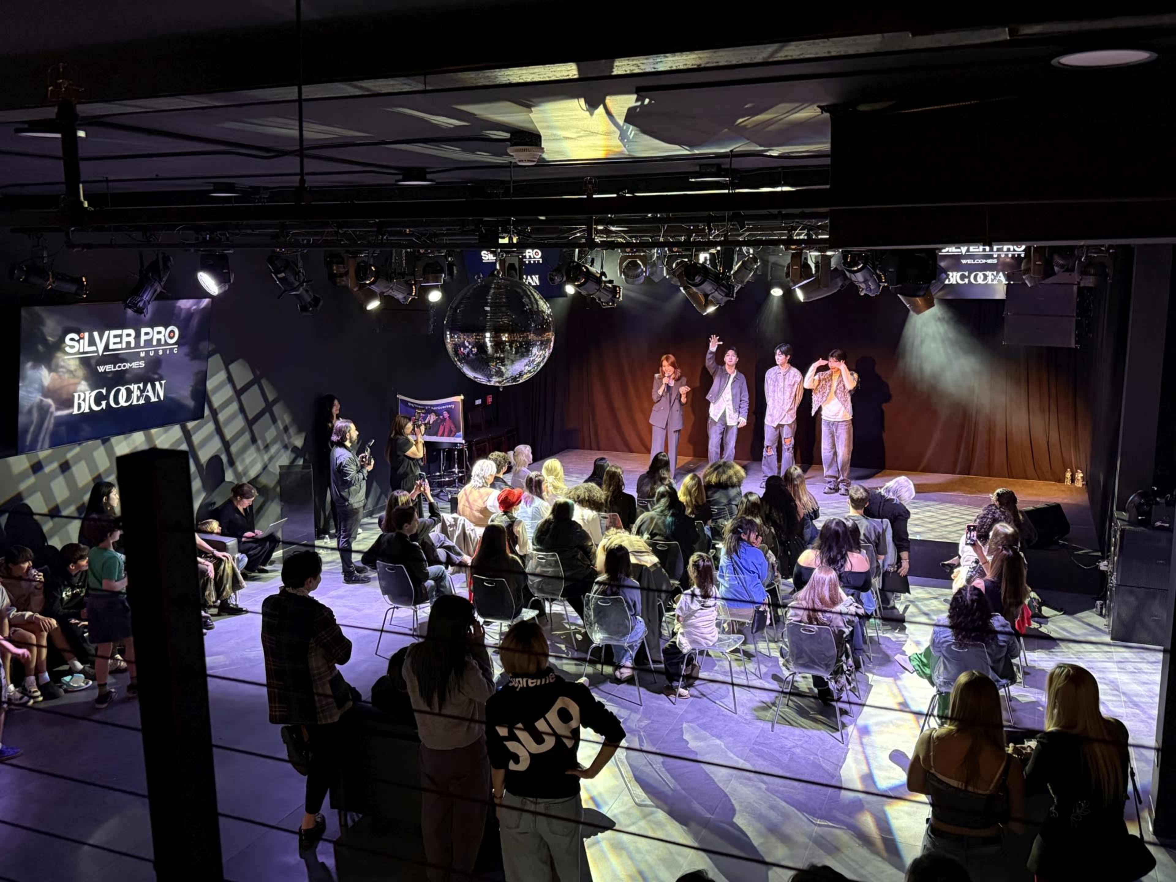 A crowd watches on as four individuals perform on stage under bright lights, with a disco ball reflecting above in a venue.