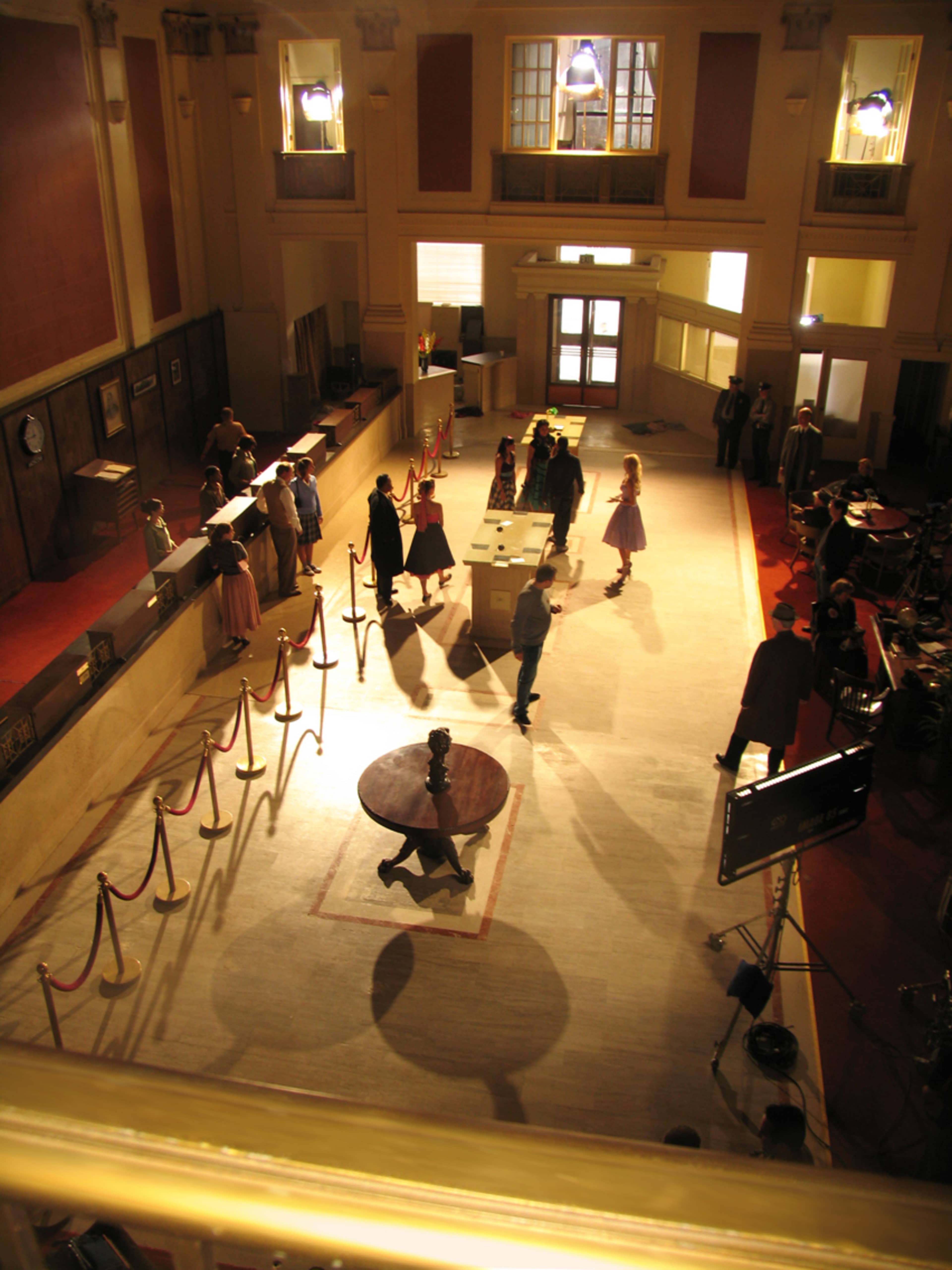 A film crew sets up a scene in a large, sunlit interior resembling a classic bank, with actors positioned near a central table and a series of lines of velvet ropes.