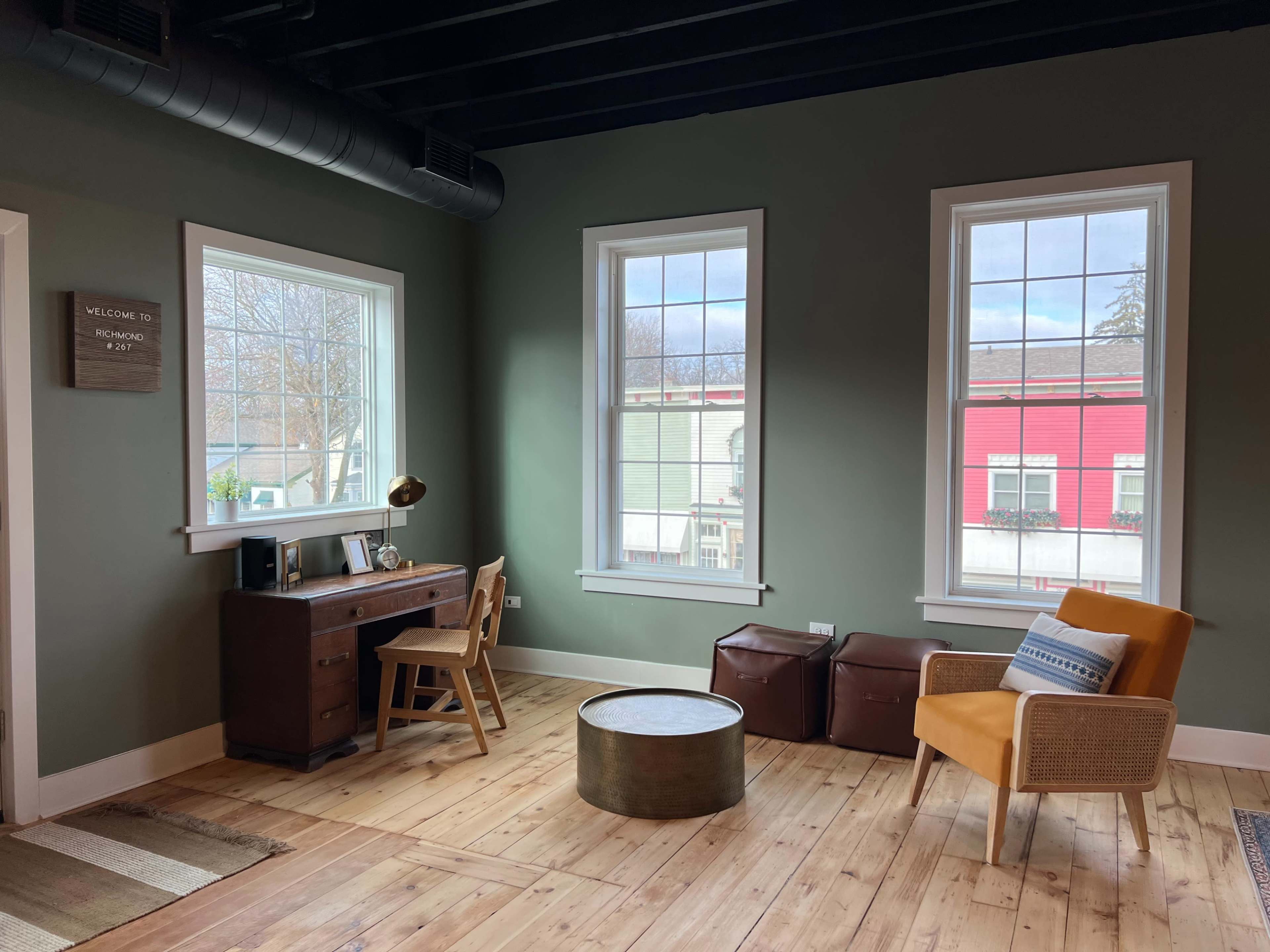 A room with green walls, two large windows, a wooden desk with a lamp, a round coffee table, and an orange accent chair.