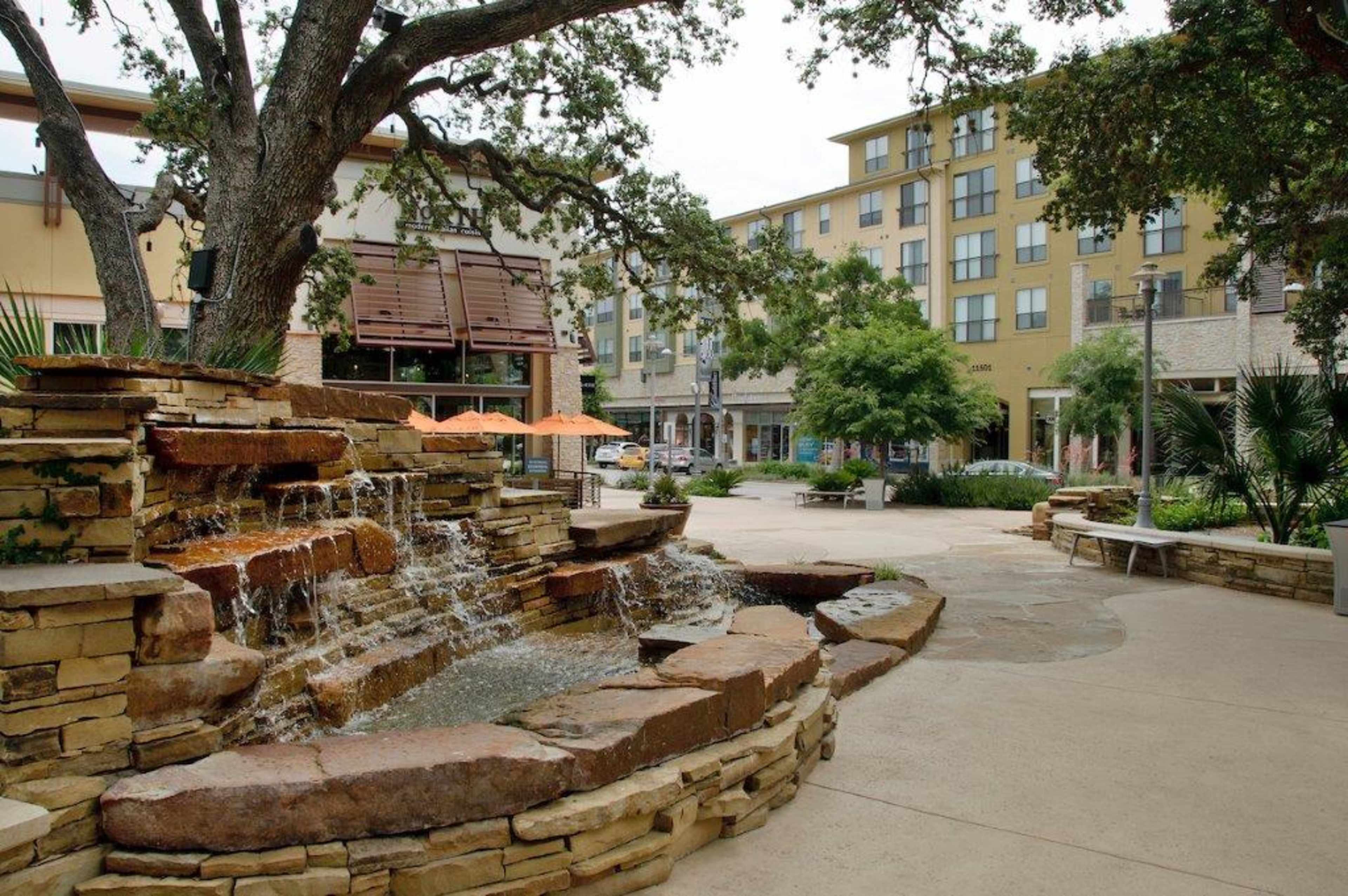 A stone water feature flows beside a walkway in an urban plaza with nearby buildings and seating areas.