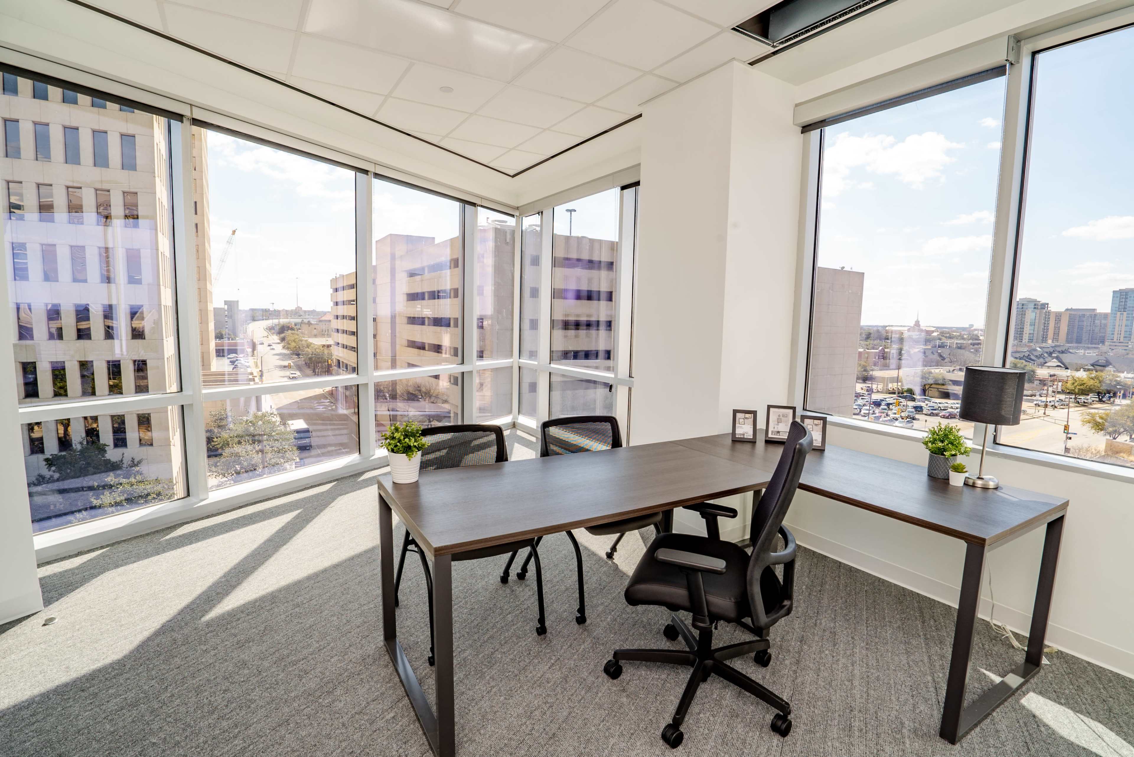A bright office space featuring a desk with two chairs, large windows that overlook a cityscape, and potted plants on the desk.