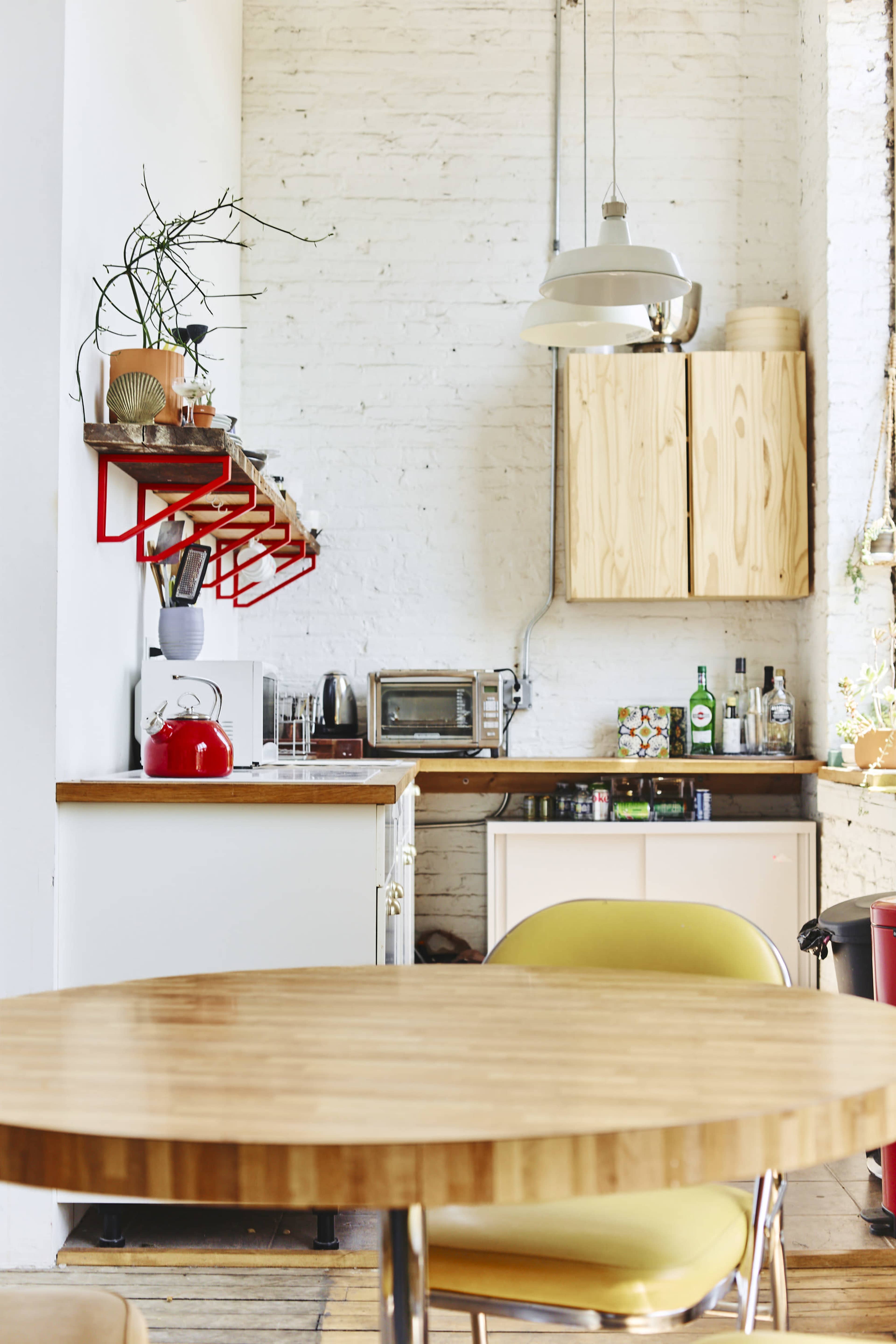 A modern kitchen features a round wooden table, open shelving with kitchen items, a red kettle, and a blend of wooden and white cabinets.