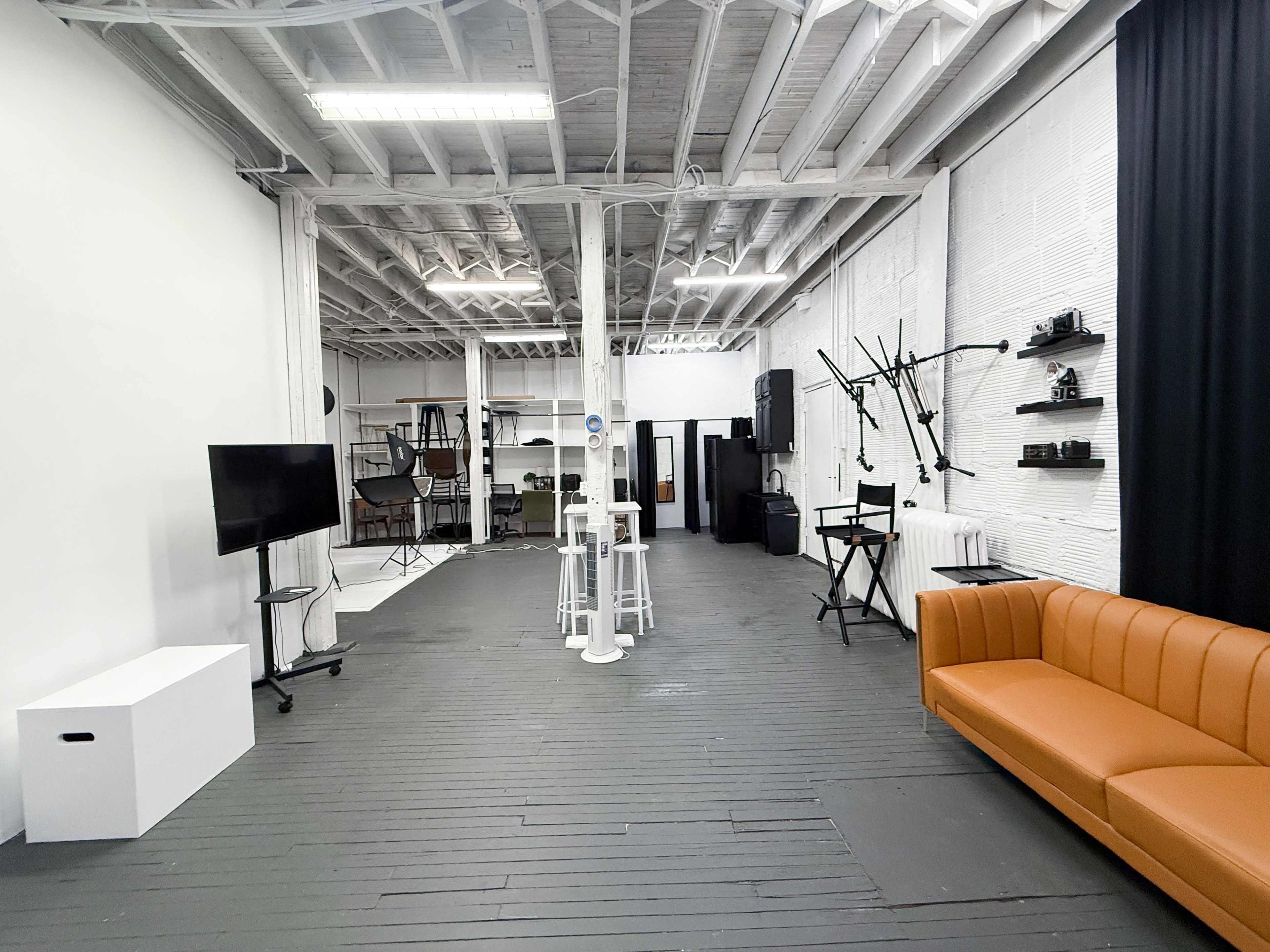 The image shows a spacious studio with wooden flooring, a brown sofa, a television on a stand, various photography equipment, and a backdrop.