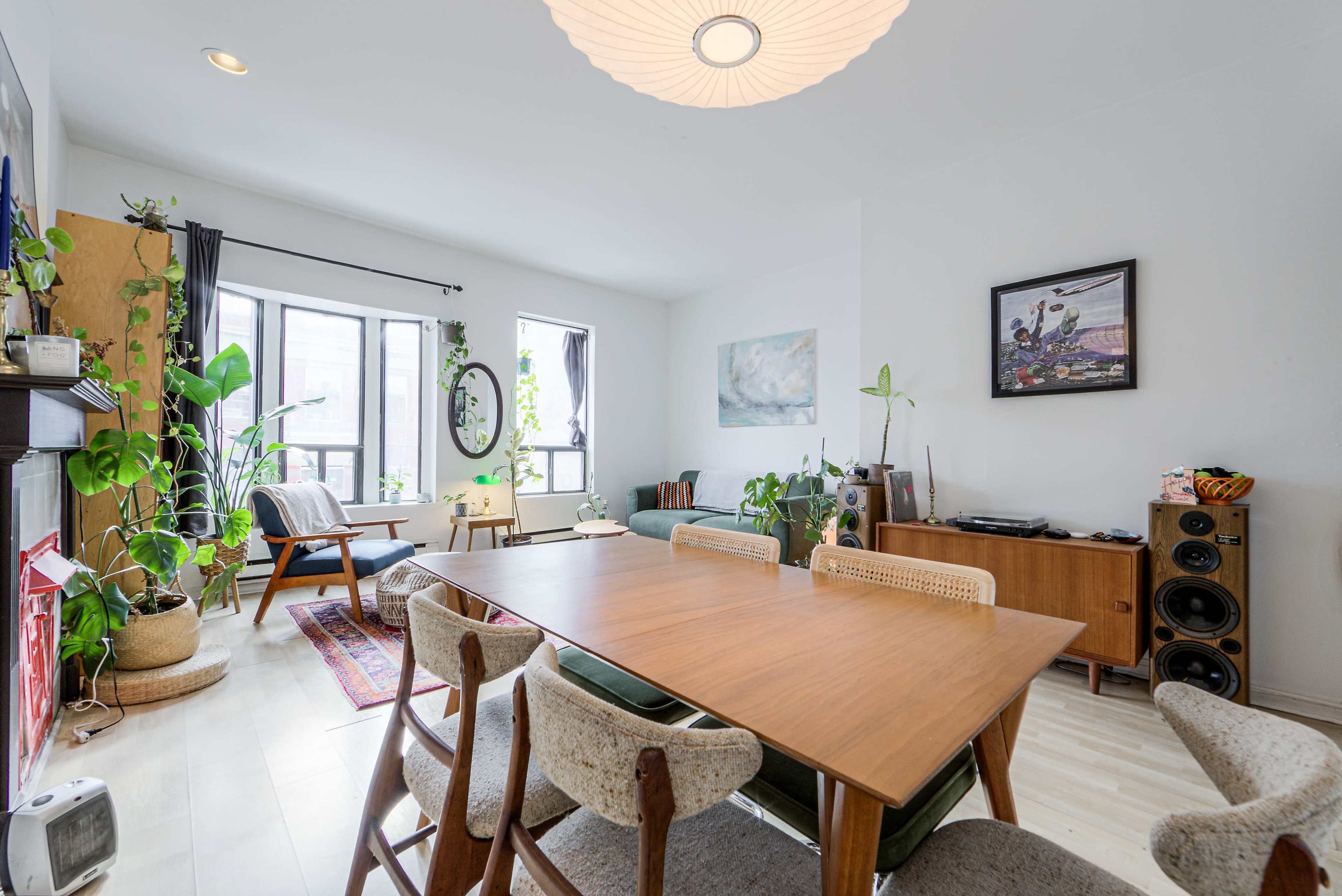 A spacious living and dining area features a large wooden table surrounded by chairs, with plants, a cozy seating area, and large windows letting in natural light.