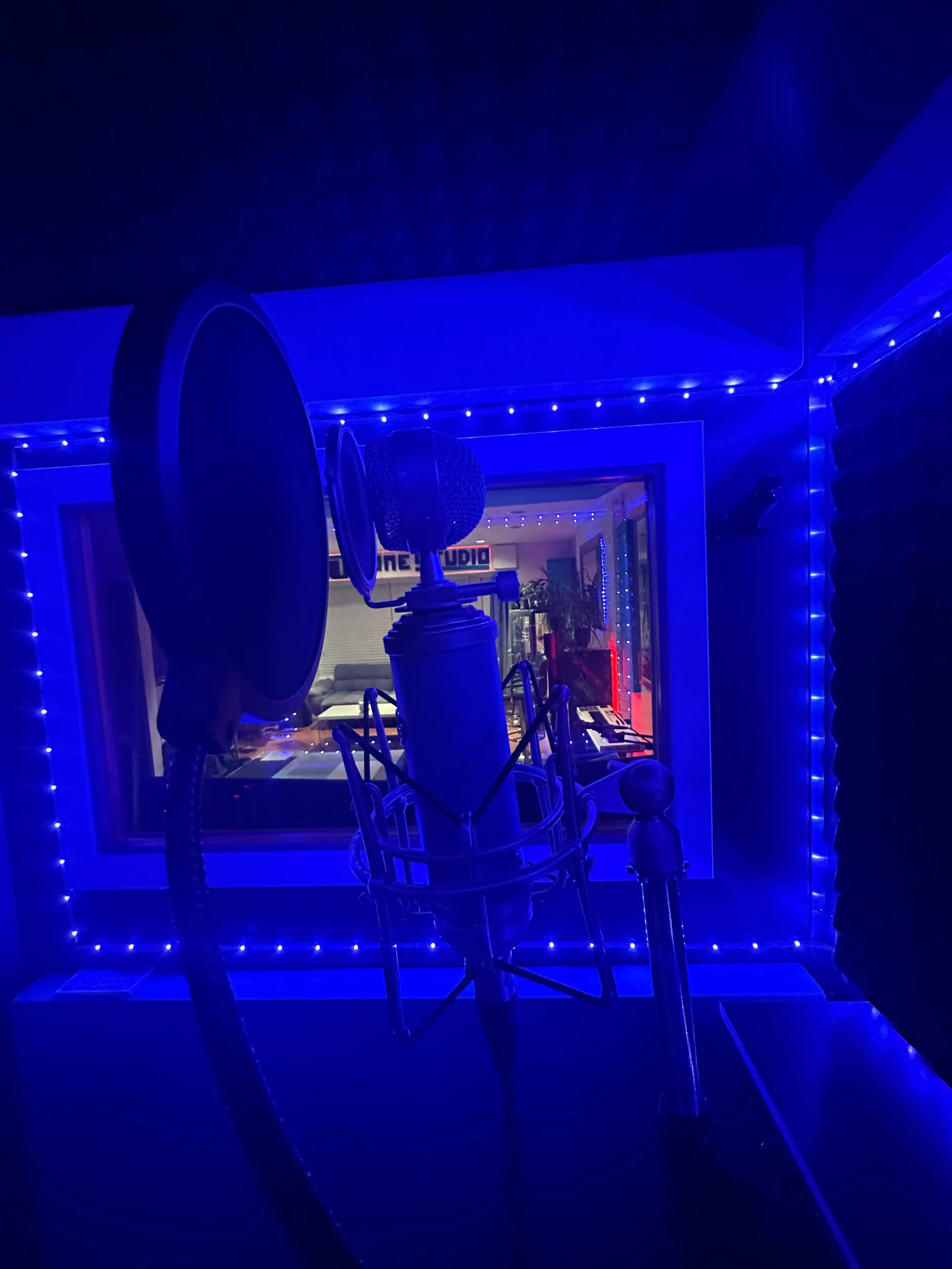 A microphone on a pop filter is positioned in front of a brightly lit recording studio with blue LED lighting.