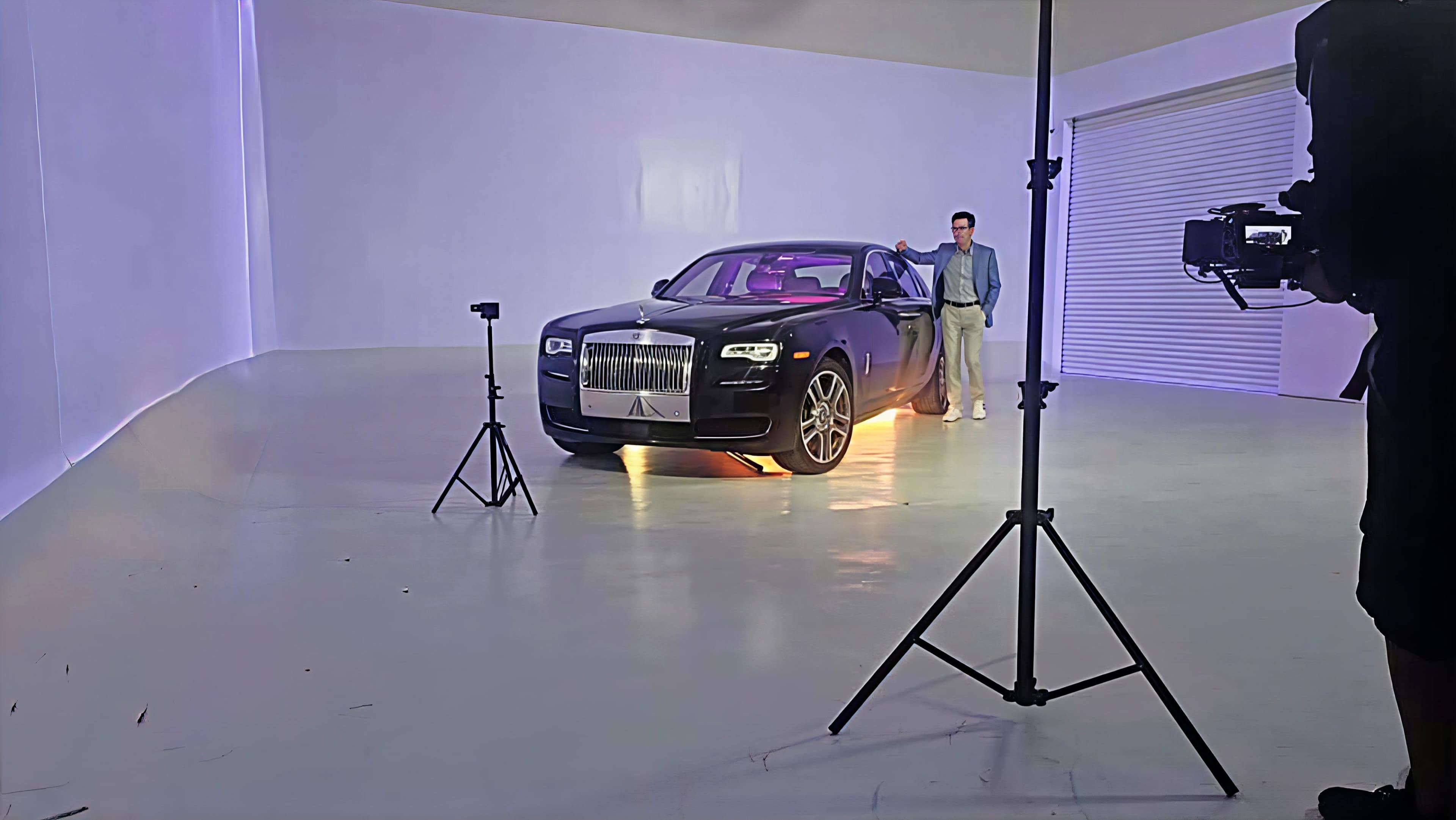 A black luxury car is positioned in an empty studio space, with a person standing next to it, while a camera on a tripod captures the scene.