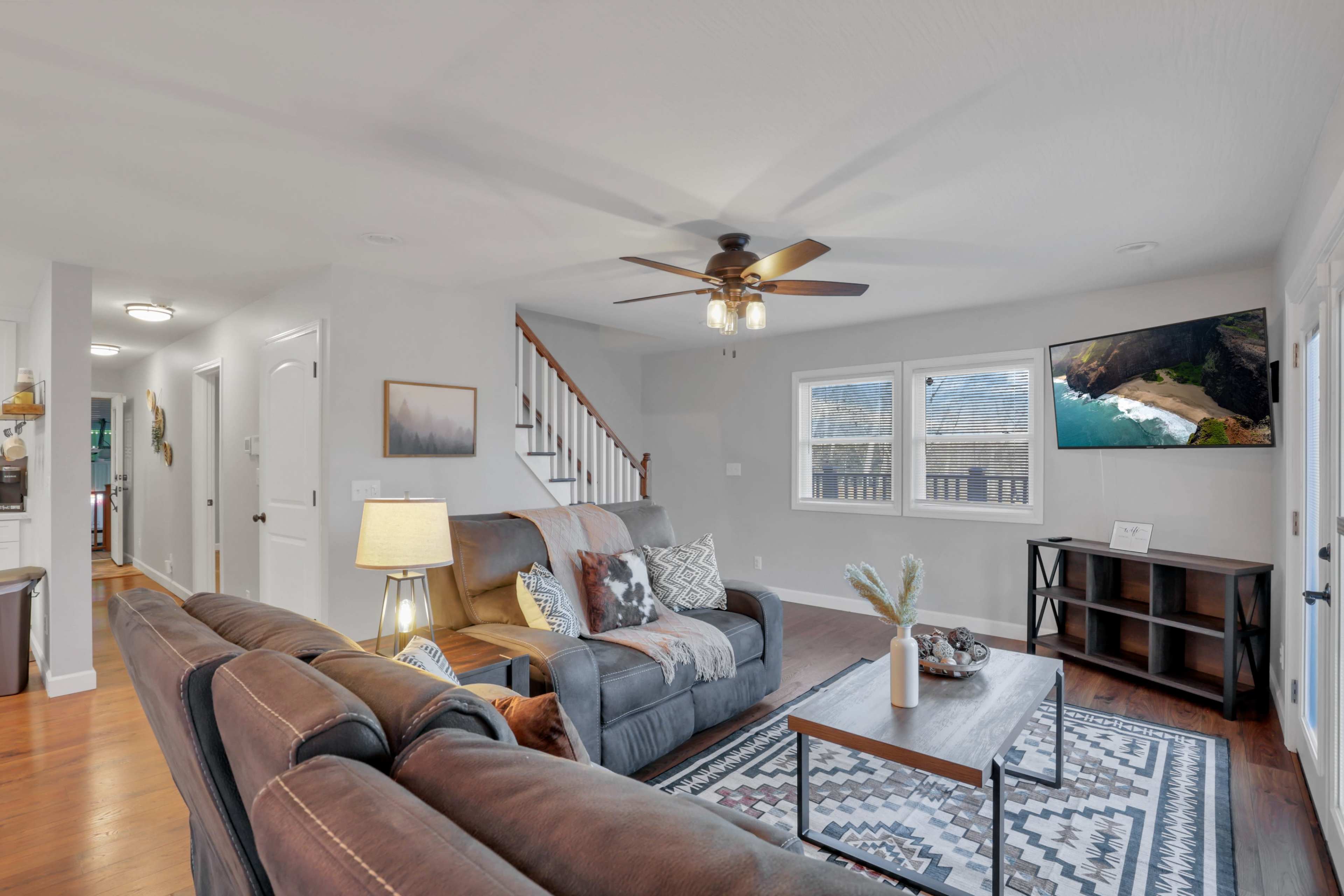 The living room features a sofa and armchair arranged around a coffee table, with a ceiling fan above and windows providing natural light.