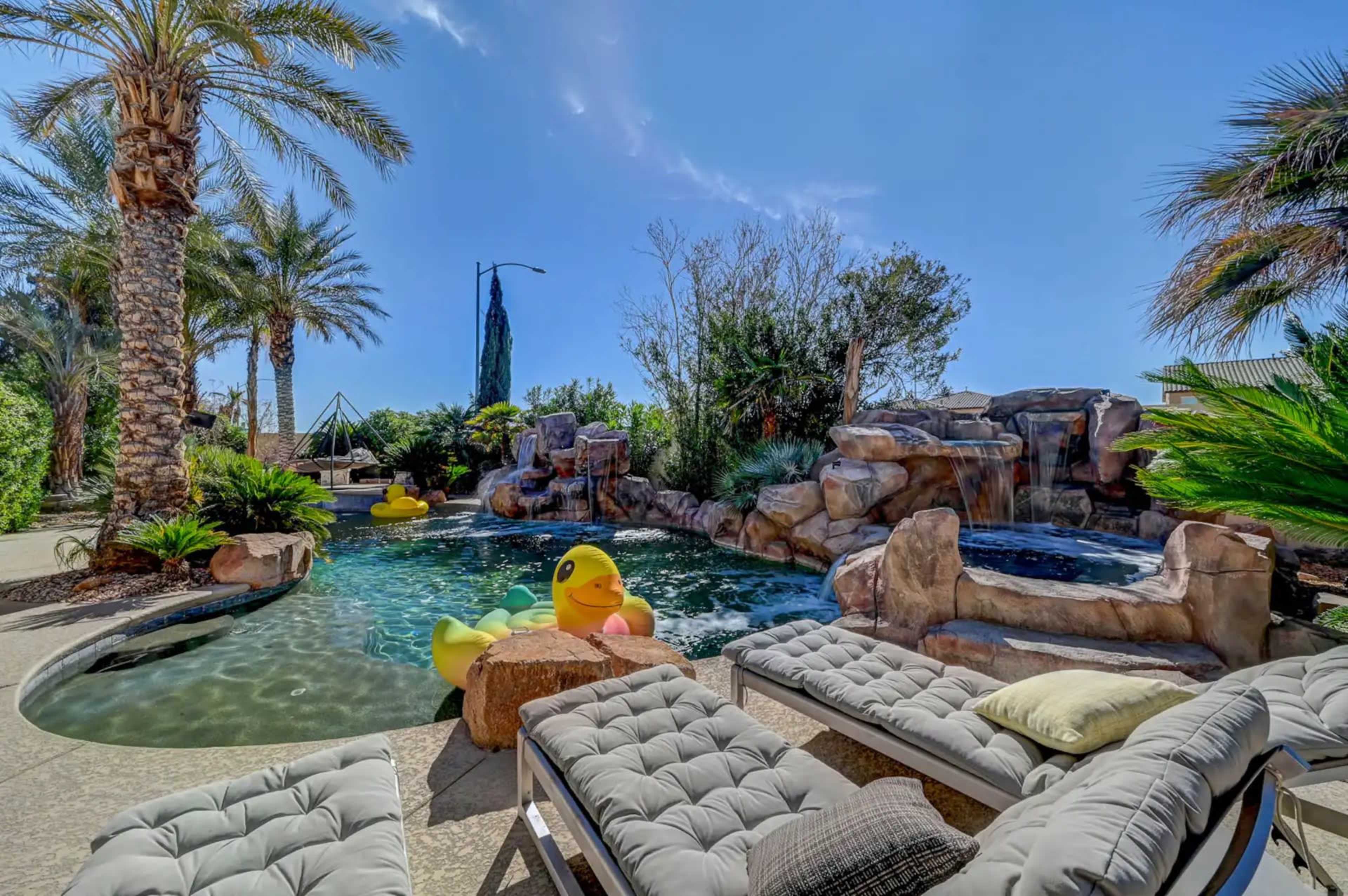 A pool area features a rock waterfall, tropical palm trees, and inflatable yellow duck floaties, with lounge chairs arranged nearby.