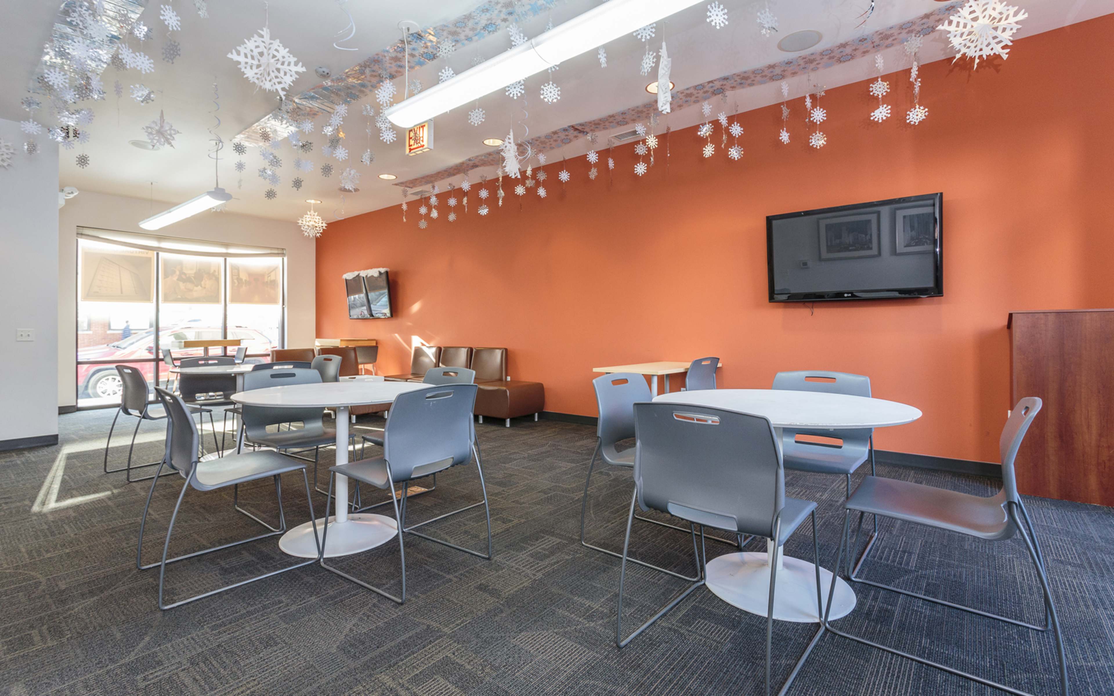 A modern community room with gray tables and chairs, an orange accent wall, and decorative snowflakes hanging from the ceiling.