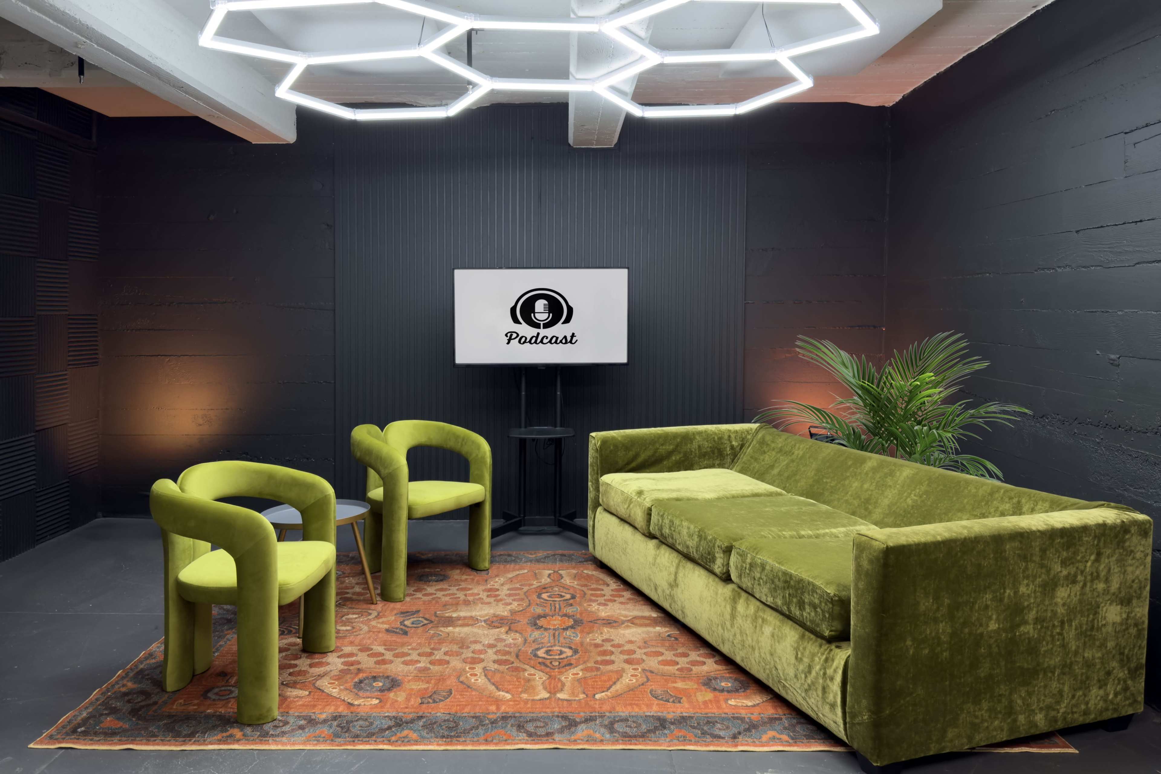 The image shows a modern podcast studio featuring a green velvet sofa, two matching green chairs, a large screen displaying the word "Podcast," and a plant, all set against a dark background.