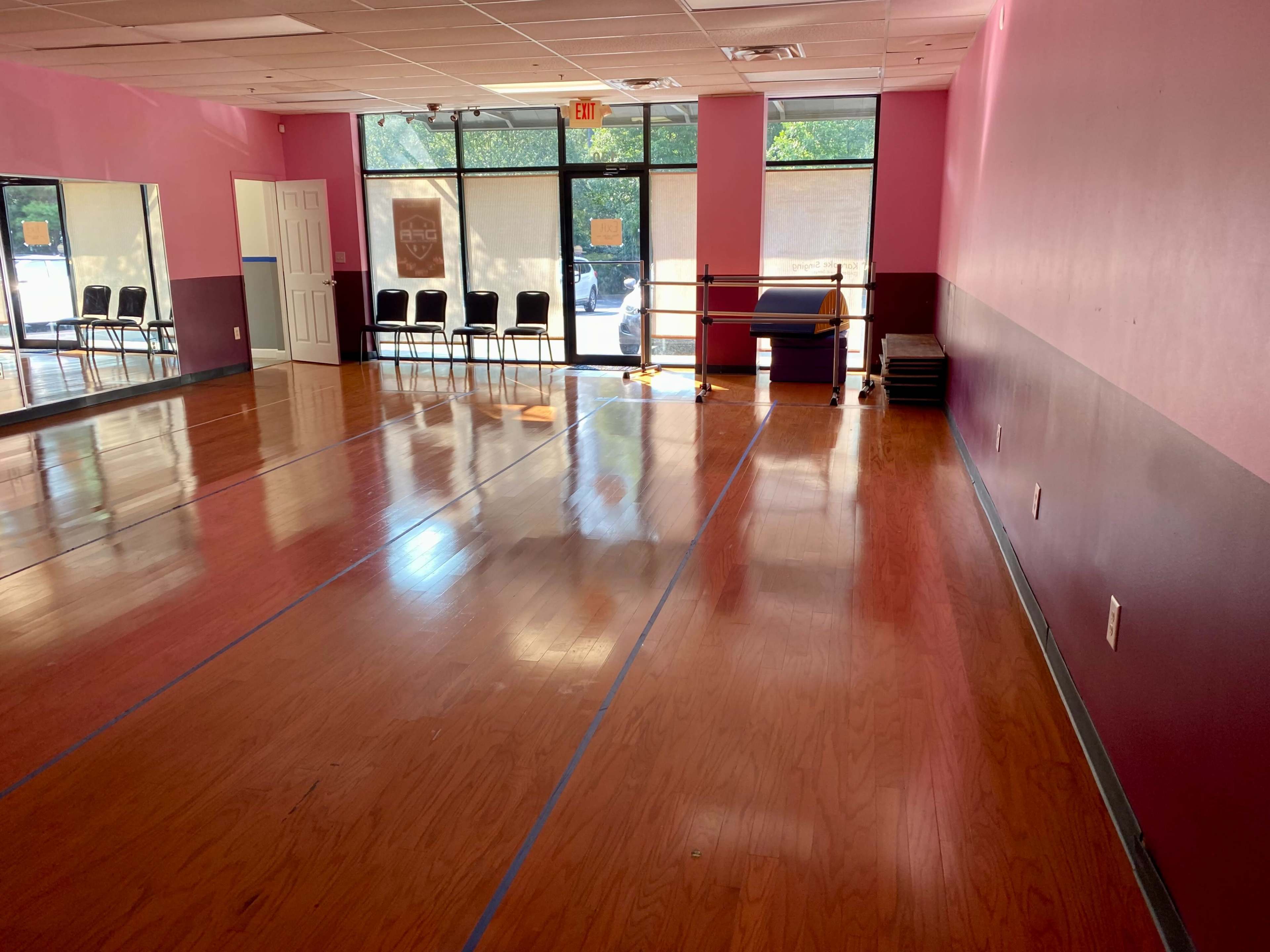 A spacious dance studio with wooden floors, pink walls, a mirrored wall, and a row of chairs near the entrance.