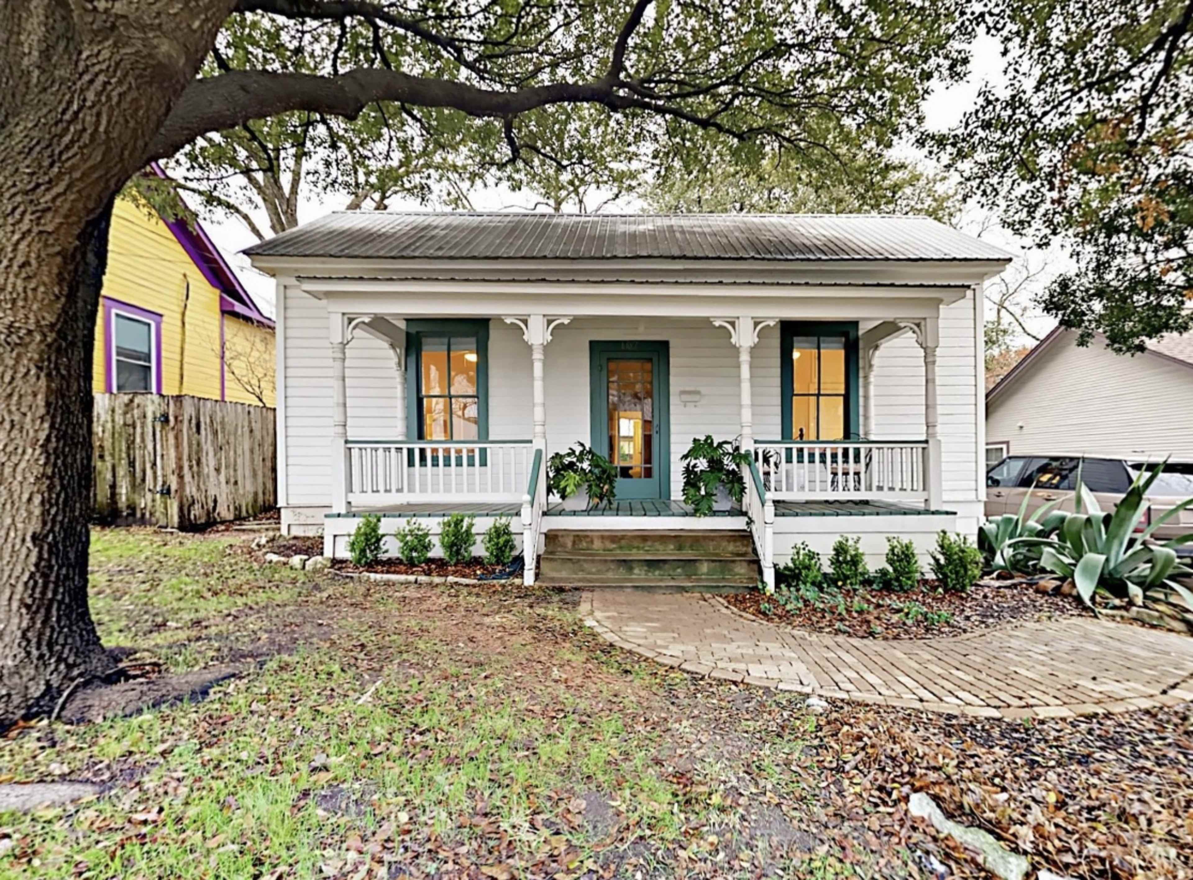 A small, white, wooden house with green shutters, a front porch, and landscaping featuring shrubs and decorative stones.