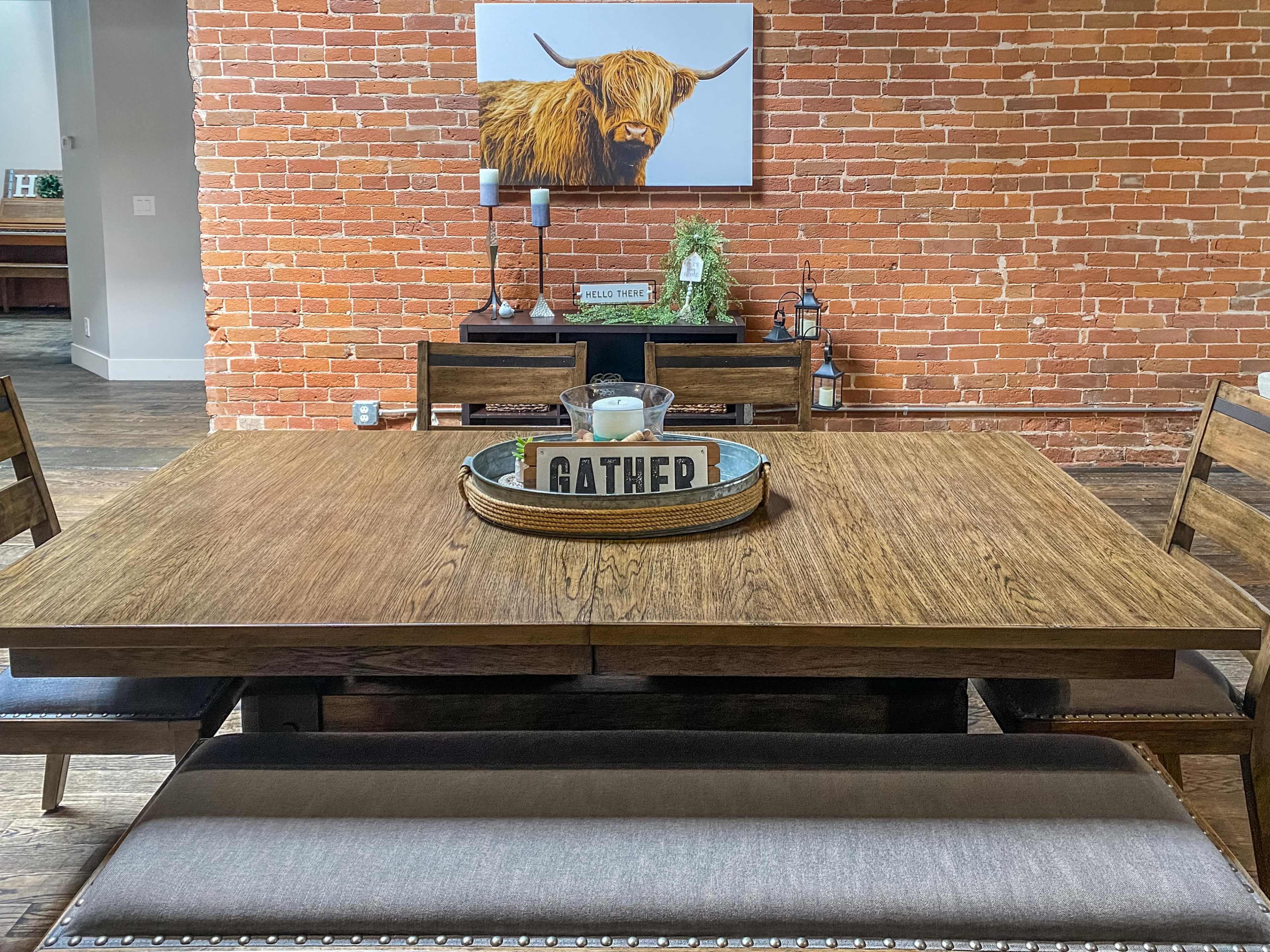 A wooden dining table with a decorative tray sits in front of a brick wall, accompanied by wooden chairs and a framed picture of a Highland cow.