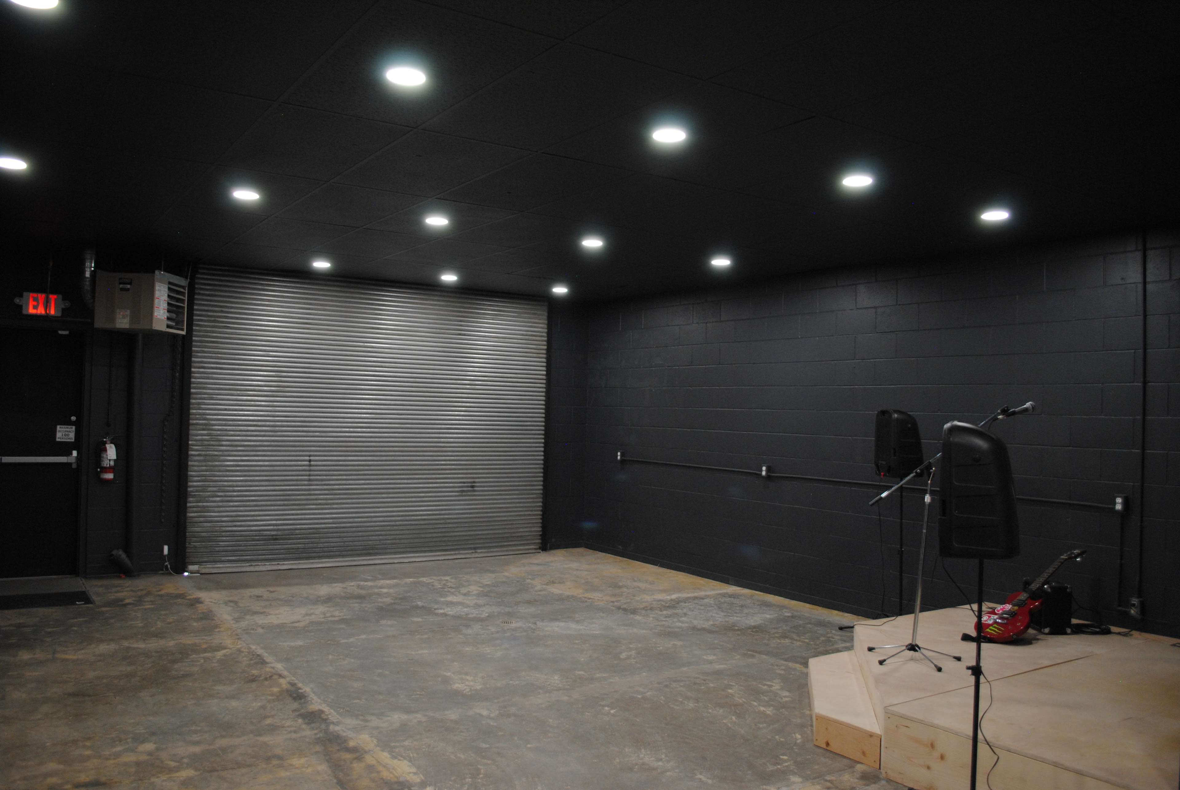 The image shows a dimly lit performance space featuring a wooden stage, a metallic roll-up door, and music equipment in the corner.