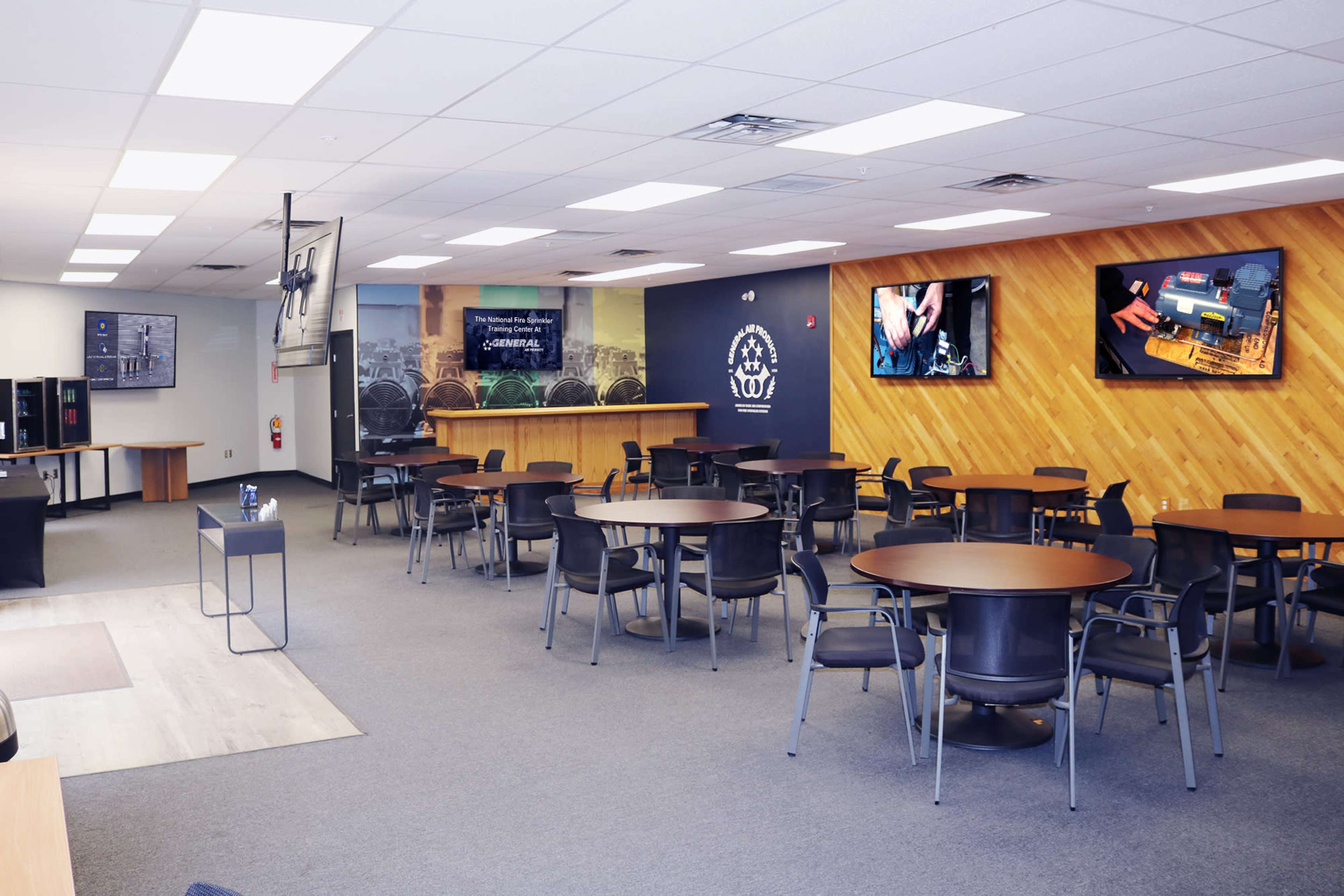 A spacious training room with multiple tables and chairs arranged for groups, along with two large screens displaying technical content on the walls.