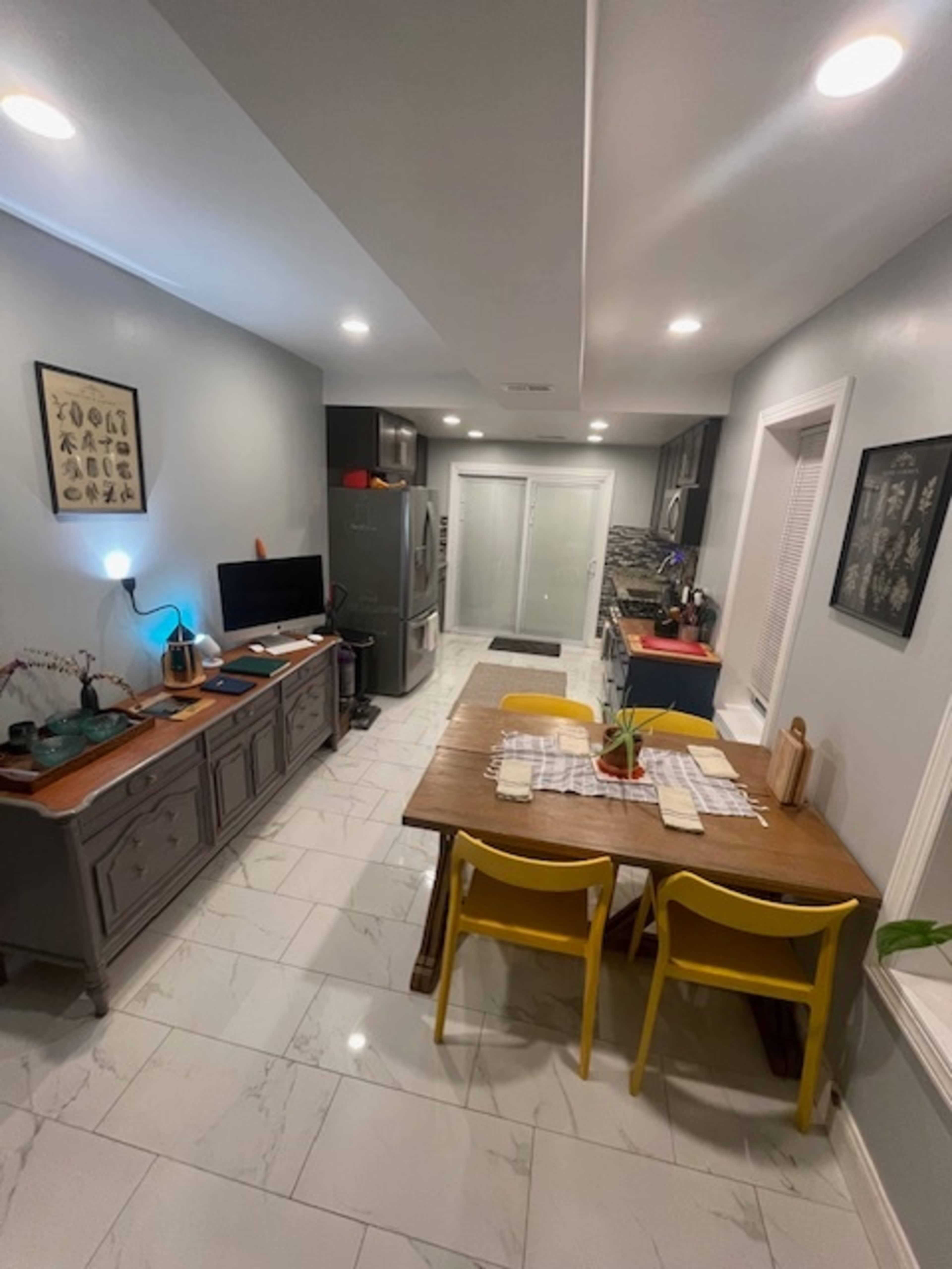 The image shows a modern kitchen featuring a stainless steel refrigerator, a wooden dining table with yellow chairs, and grey cabinets against tiled flooring.