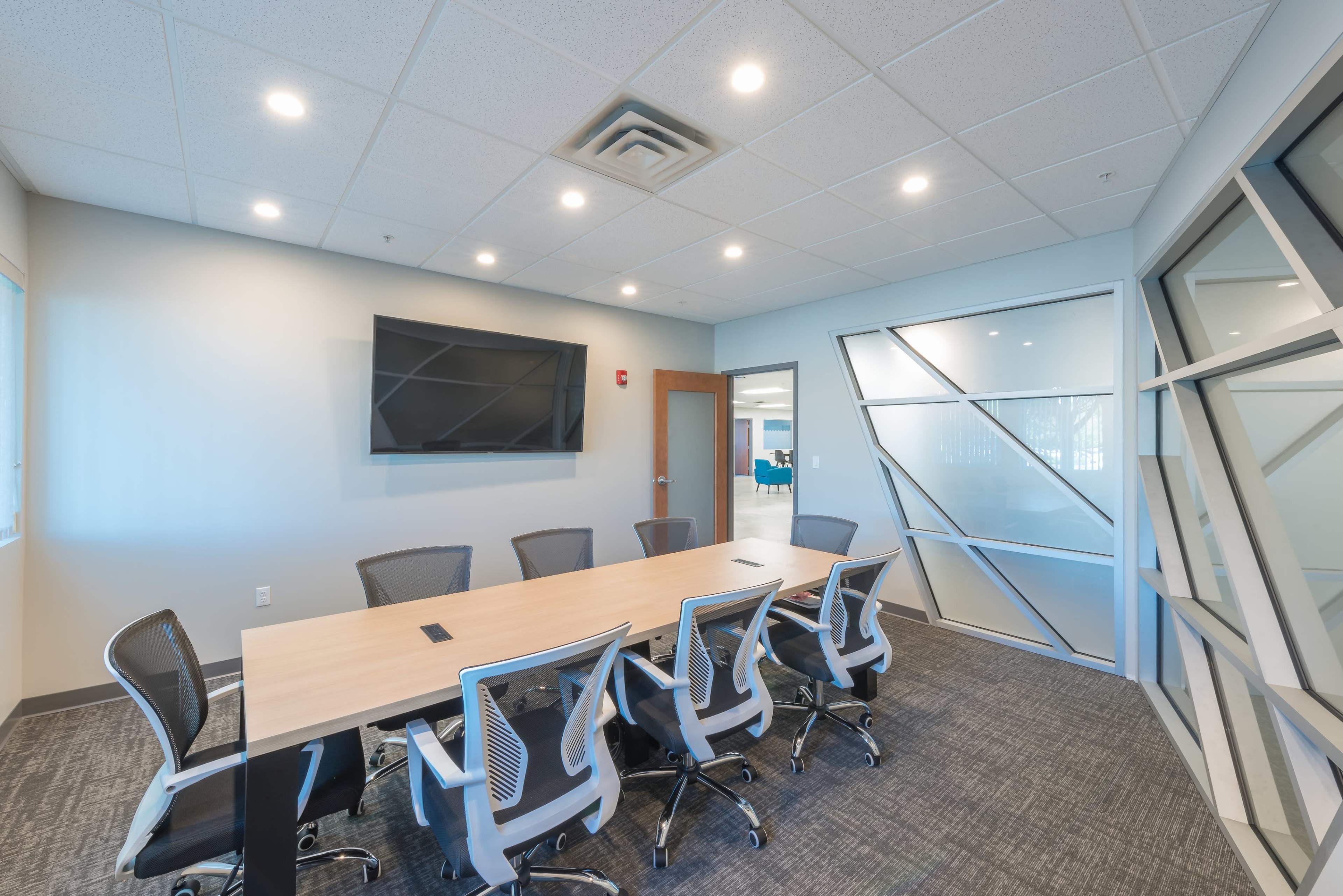 A modern conference room features a large table surrounded by ergonomic chairs, with a wall-mounted television and large windows allowing natural light.
