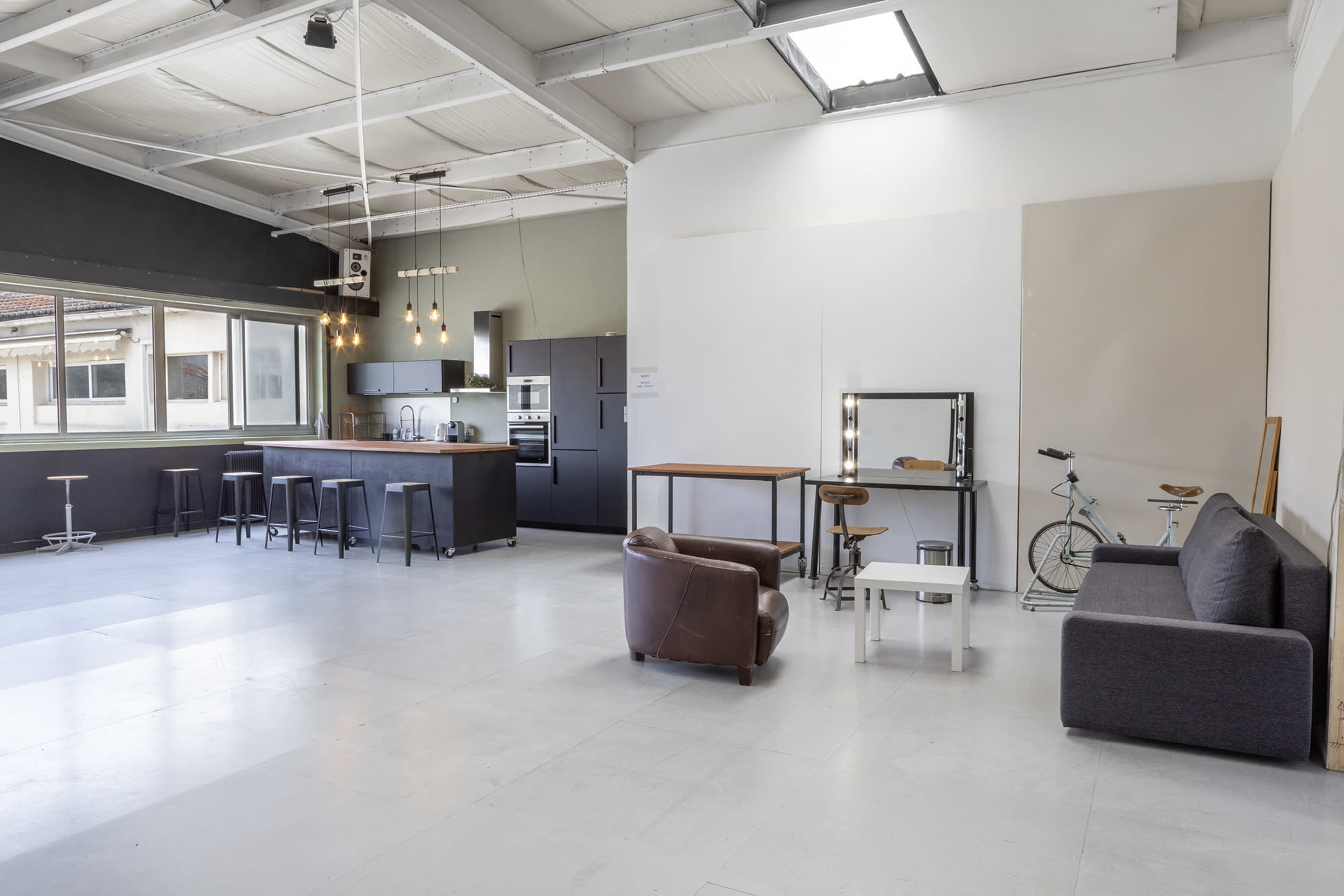 The image shows a spacious, modern interior with a kitchenette, seating area, and a bicycle propped against the wall.